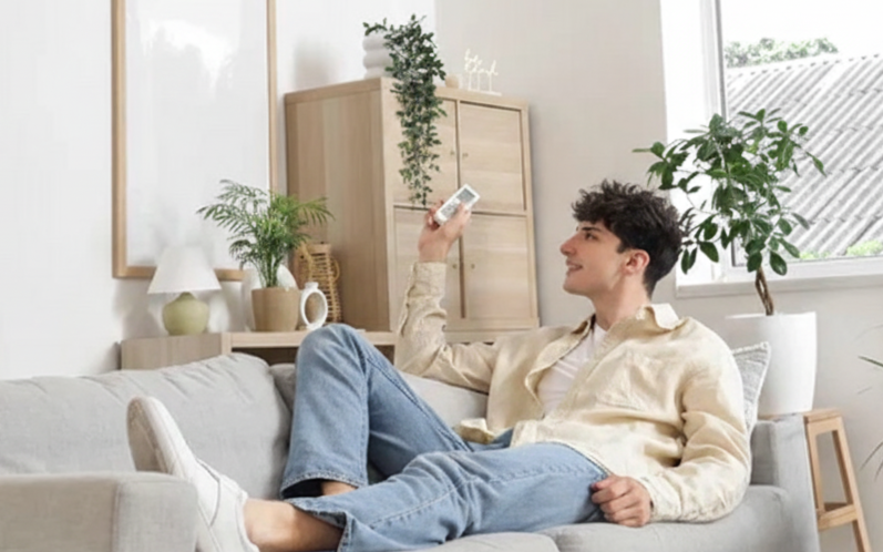 Junger Mann liegt entspannt auf einem Sofa, hält eine Fernbedienung in der Hand und schaut freundlich.