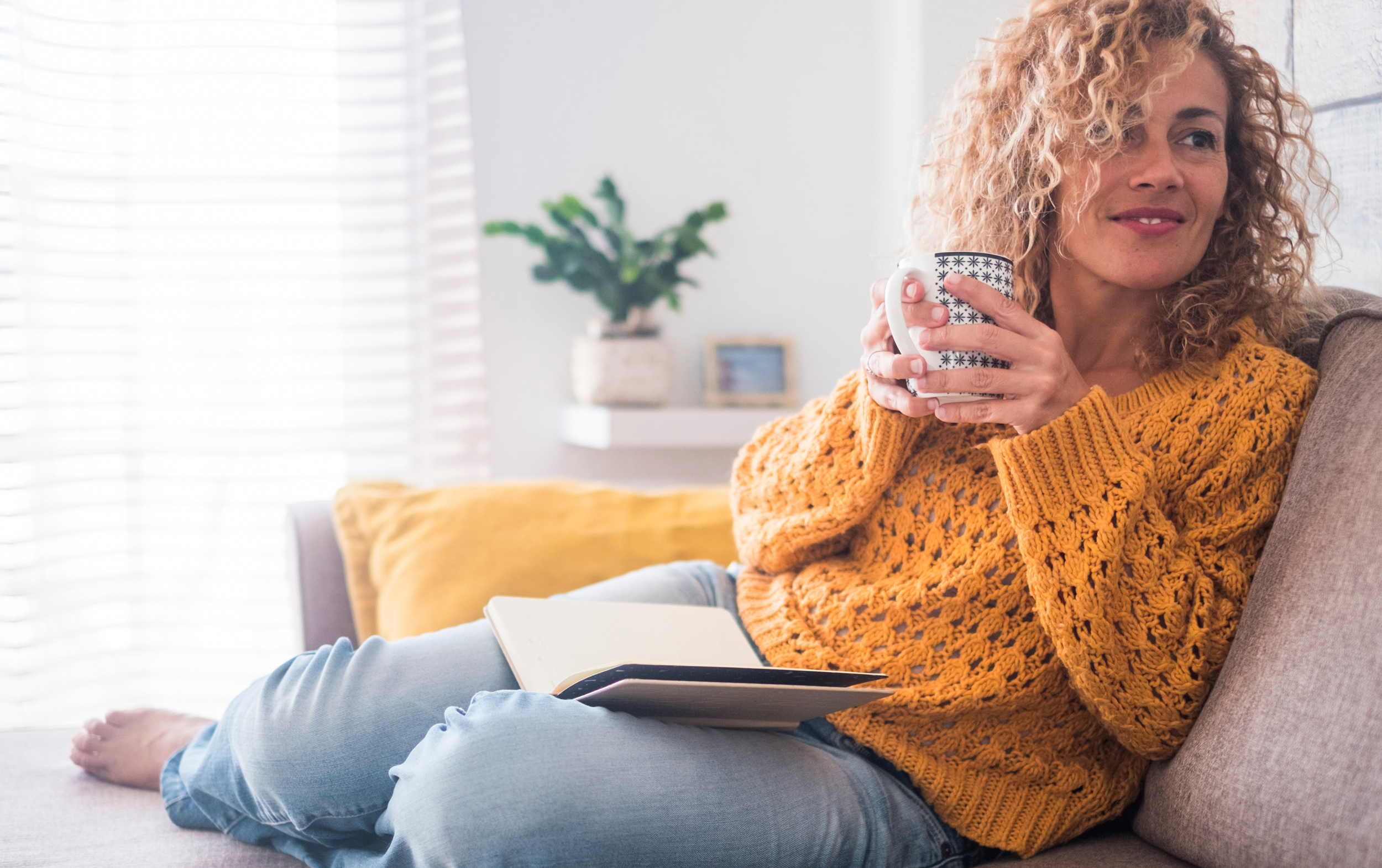 Frau mit lockigem Haar sitzt auf einem Sofa, hält eine Tasse und liest ein Buch, während sie sich entspannt. Sie trägt einen gelben Strickpullover.