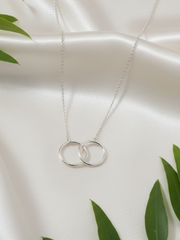 Silver necklace with two interlocking rings pendant on white satin fabric, with green leaves in the foreground.