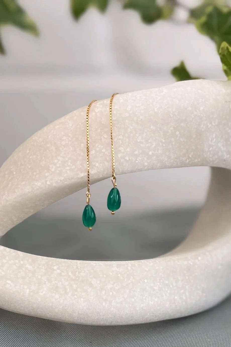 Gold chain earrings with green teardrop-shaped stones hanging from a white textured display piece with green leaves in the background.