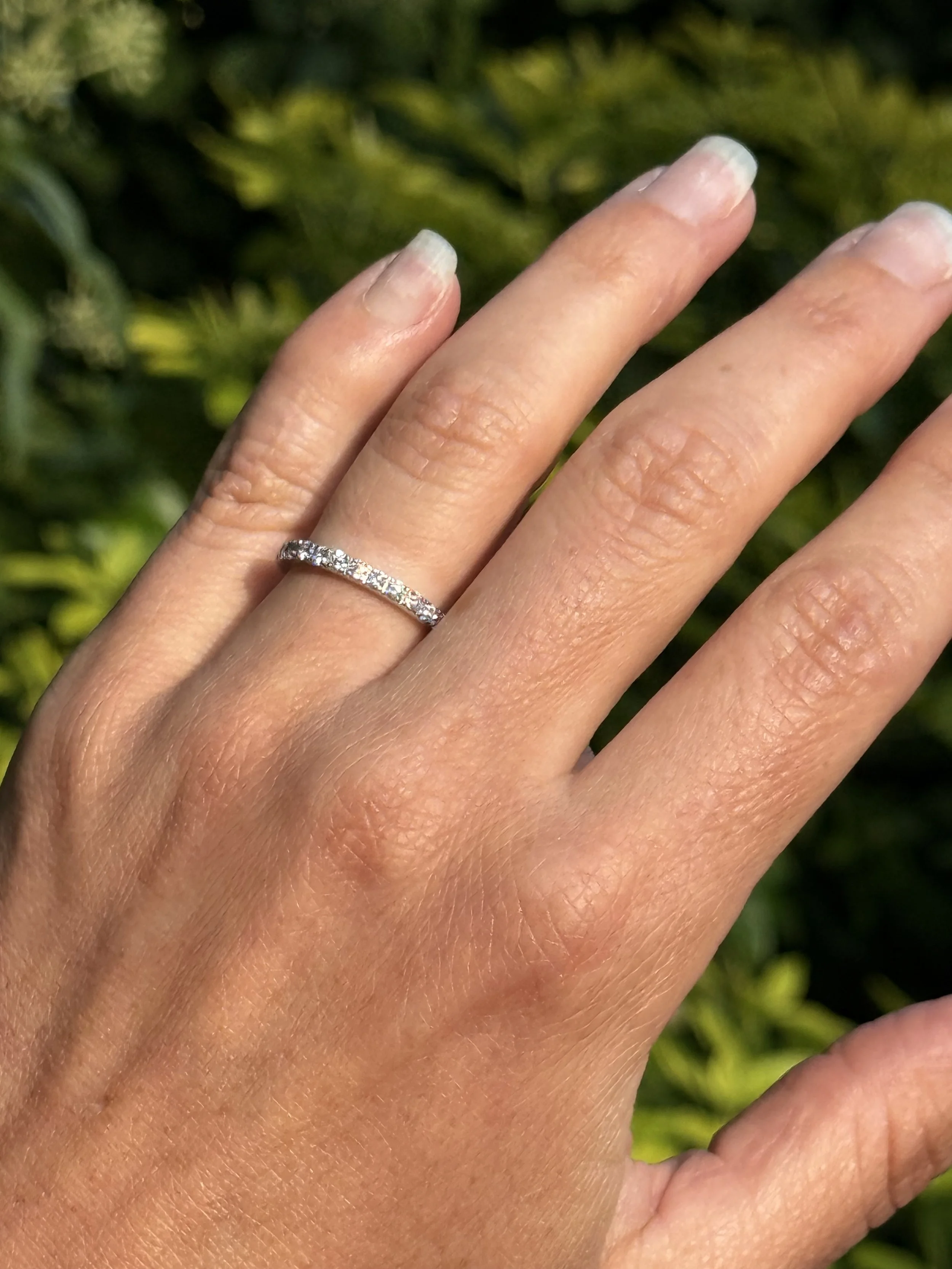 A close-up of a woman's left hand showing a diamond engagement ring on her ring finger, with a blurred green background.