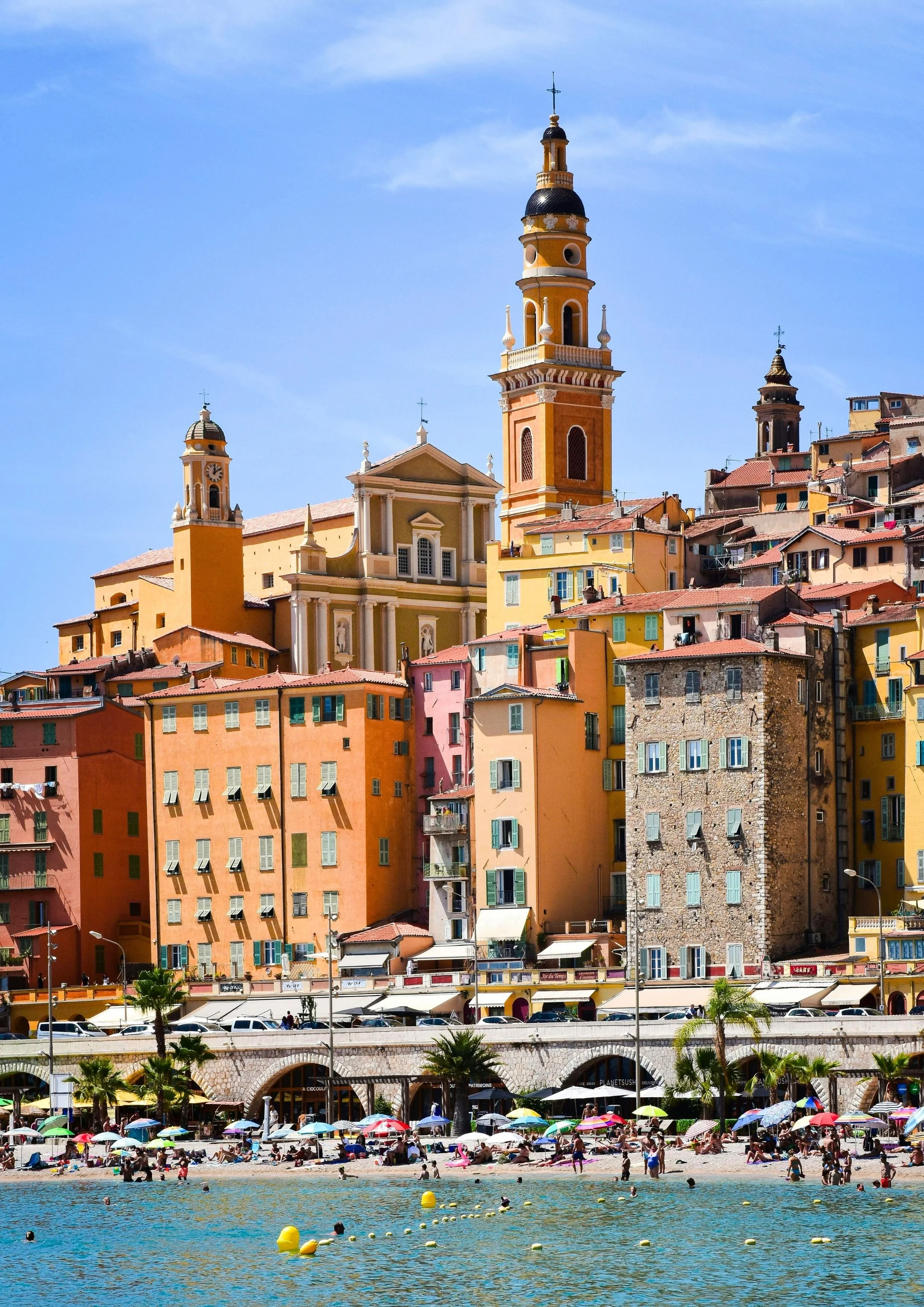 Coastal scene with rocky shoreline, trees, and hillside houses overlooking the water on a clear day.