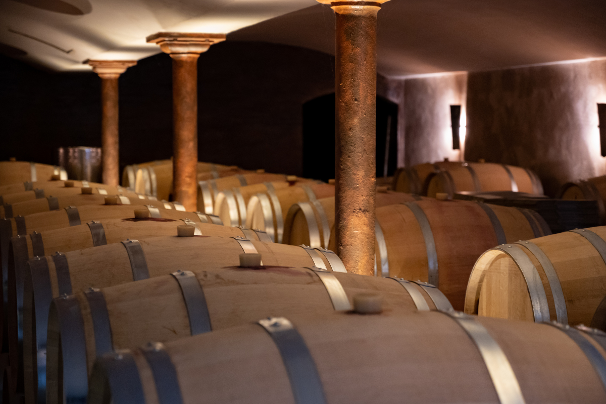 Rows of wooden wine barrels stored in a cellar with metal hoops, supported by rust-colored pillars, and dim lighting.
