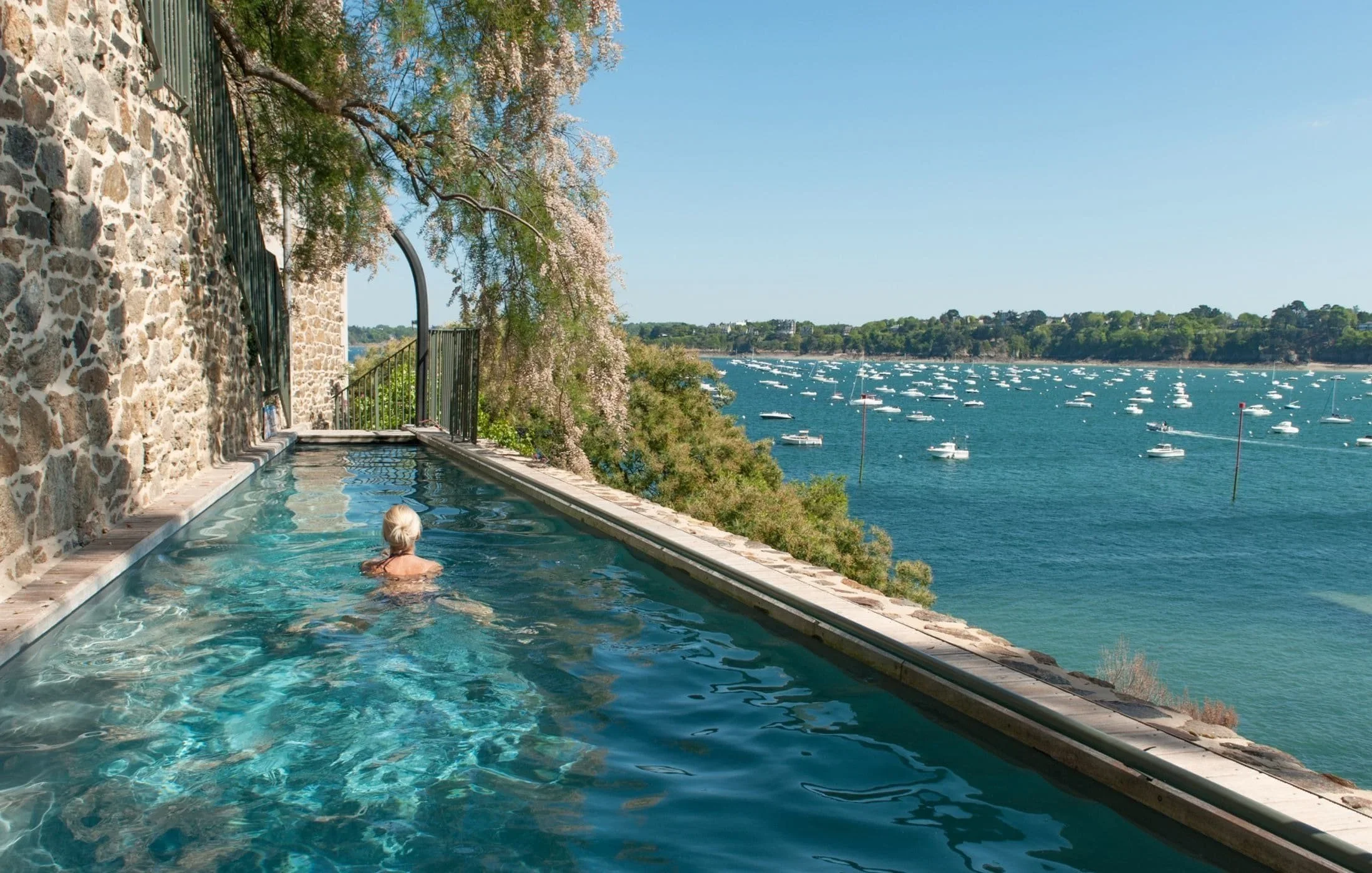 Elderly person swimming in an elevated pool overlooking a sea filled with boats and a green landscape in the background.