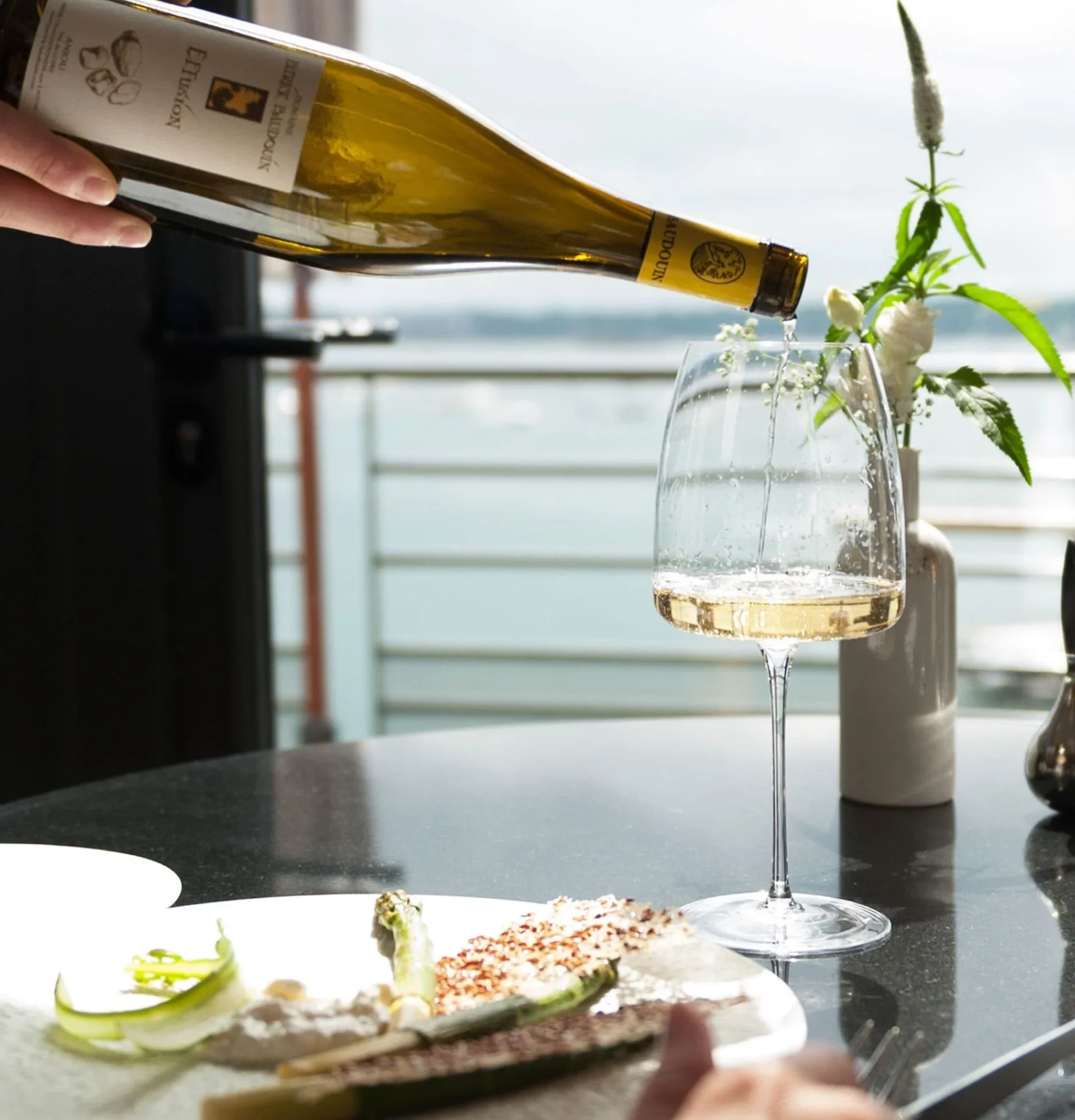 A hand pours white wine into a wine glass on a table with a bottle of wine, a plate of food, and a vase of flowers, in front of a balcony overlooking the water and sky.