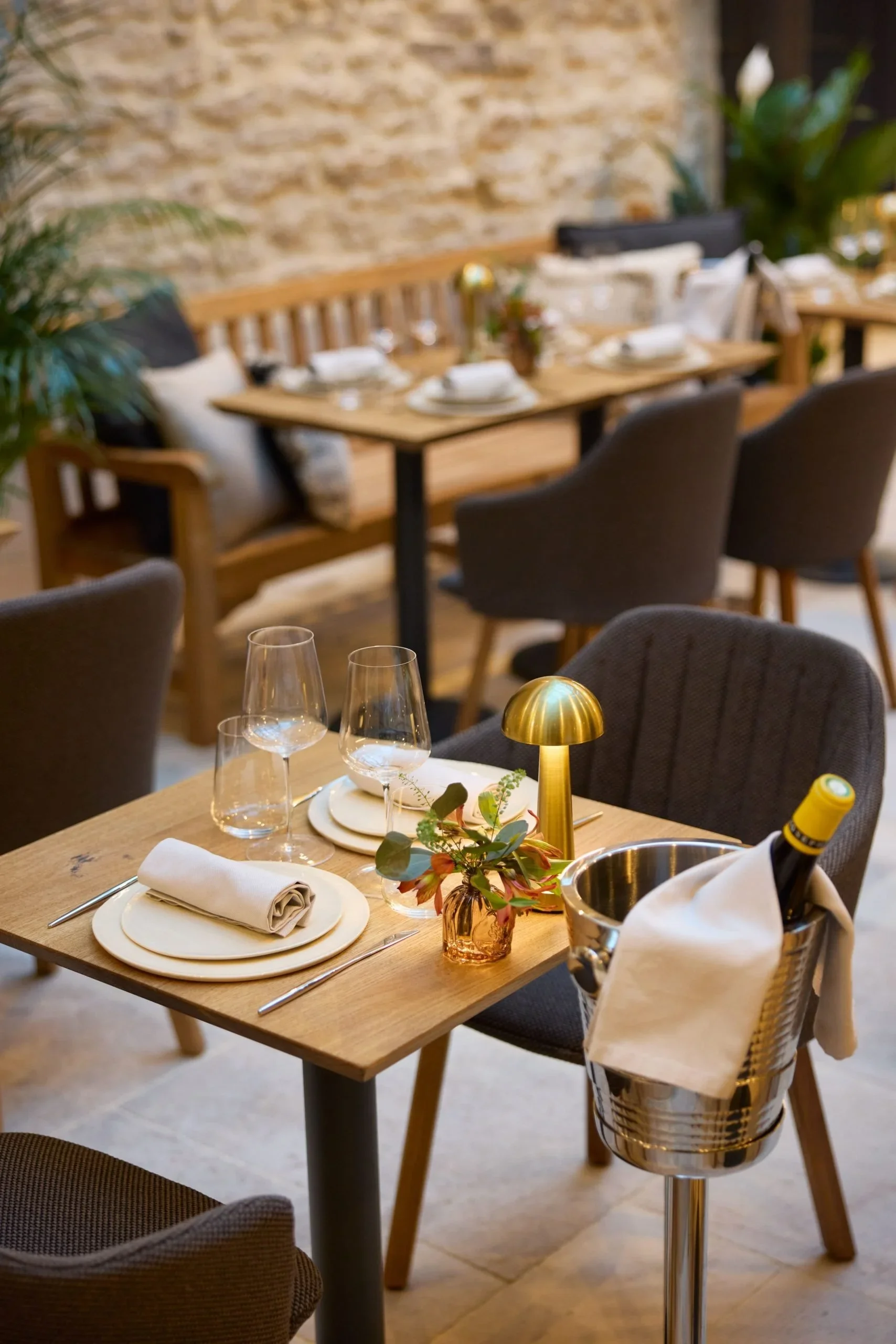 Table de restaurant élégante avec vaisselle, verres, une bouteille de vin dans un seau à champagne, décoration florale et lampe dorée, dans un cadre chaleureux avec murs en briques