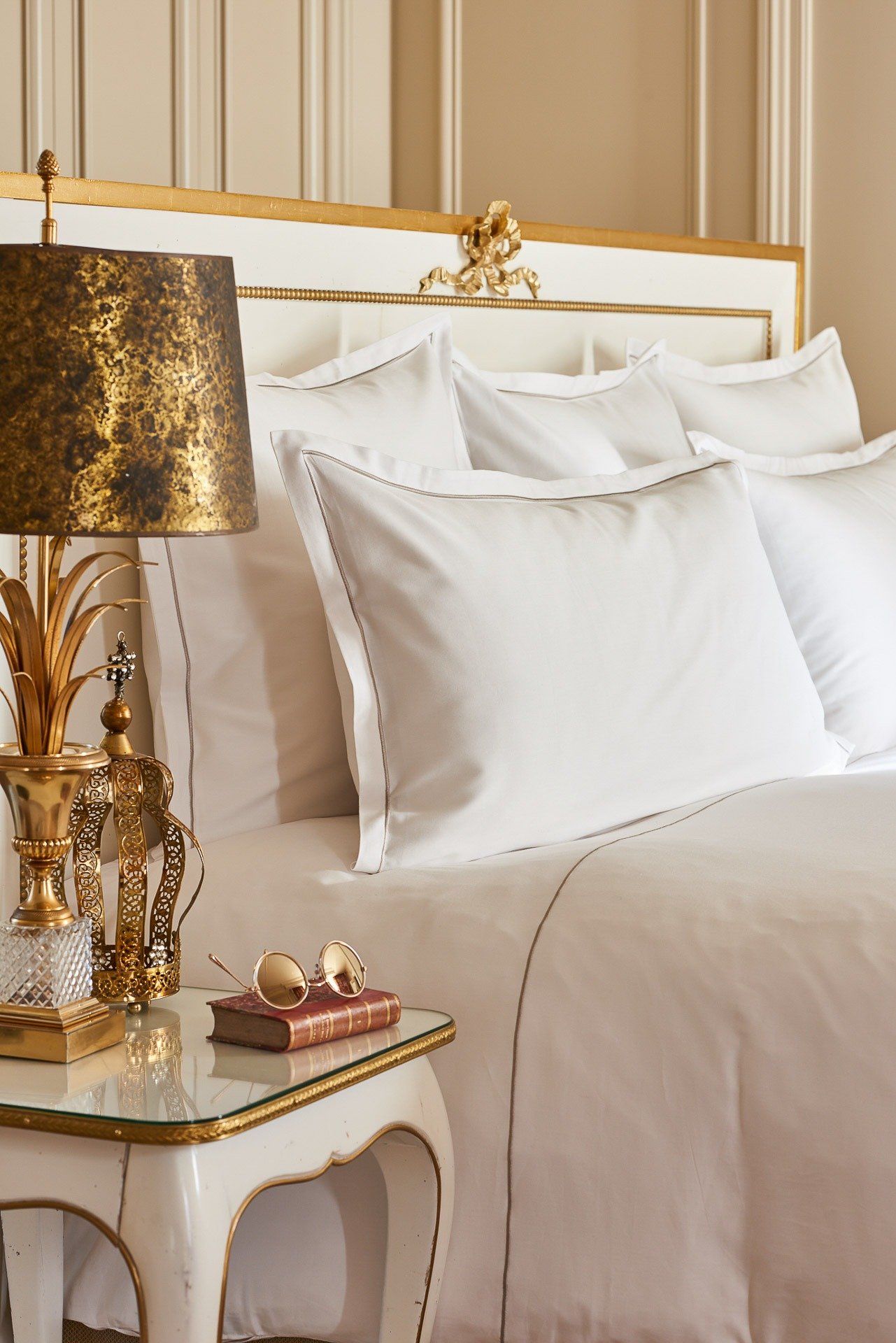 Un lit avec plusieurs oreillers blancs, une table de chevet avec des lunettes de soleil, un livre et une lampe dorée