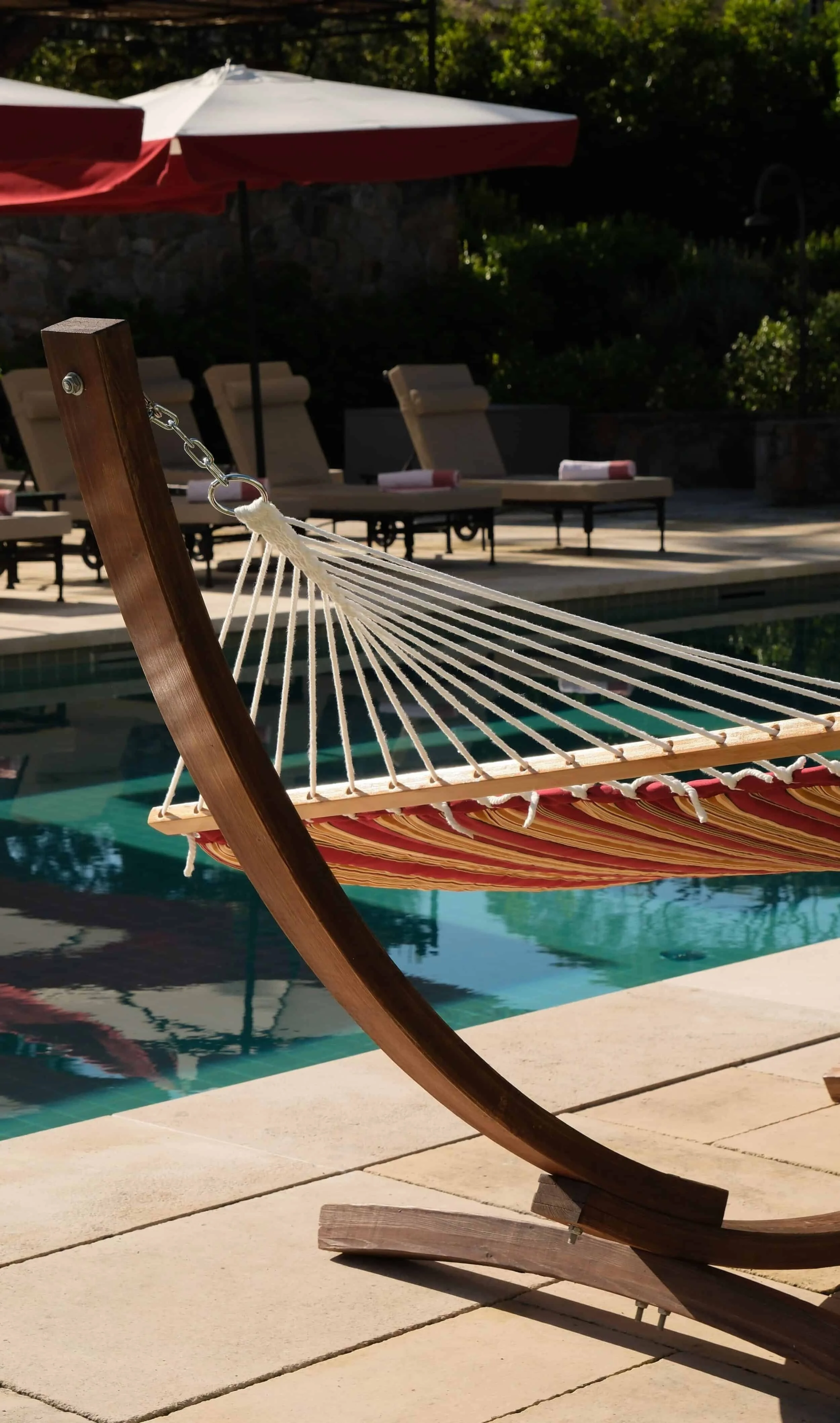 Wooden hammock with red and beige striped fabric, located near a swimming pool in an outdoor area with lounge chairs and a red and white parasol.