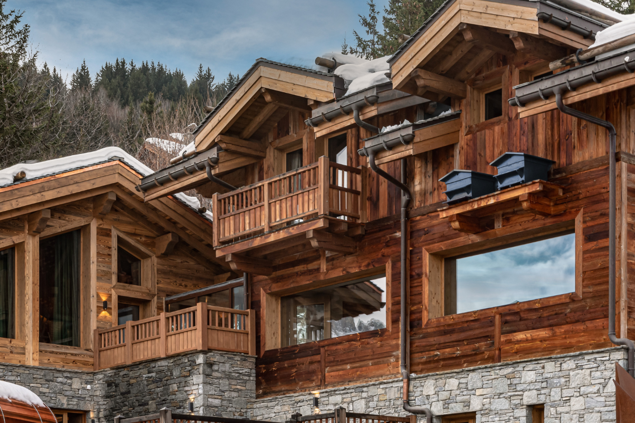 View of a wooden house built on a hill, with snow-covered roofs, surrounded by trees, under a cloudy sky.