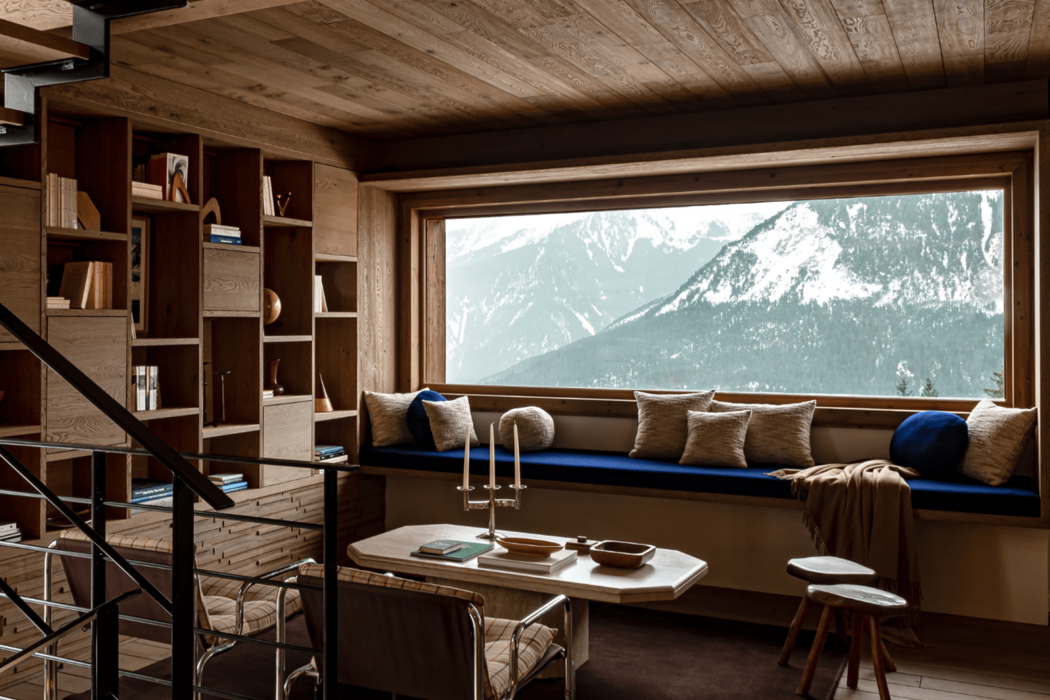 Living room with a view of a snow-capped mountain, wooden bookcase, built-in sofa with several cushions, coffee table with books and candles, metal armchairs, prefabricated metal and wood staircase.