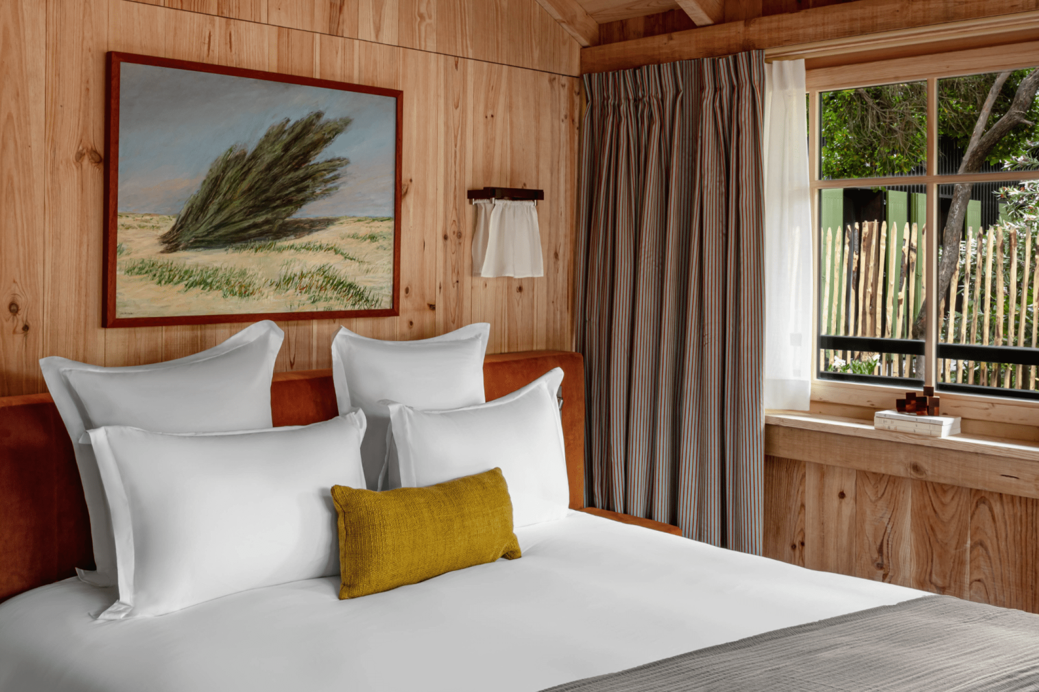 Bedroom with several white pillows and a yellow cushion, wooden walls, landscape painting, window with curtains and a view of a garden.