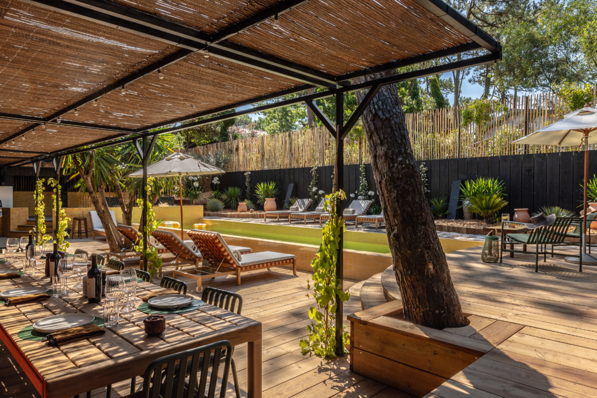An outdoor relaxation area with a swimming pool, lounge chairs, umbrellas, and a dining table under a shelter. Green surroundings with trees and plants.