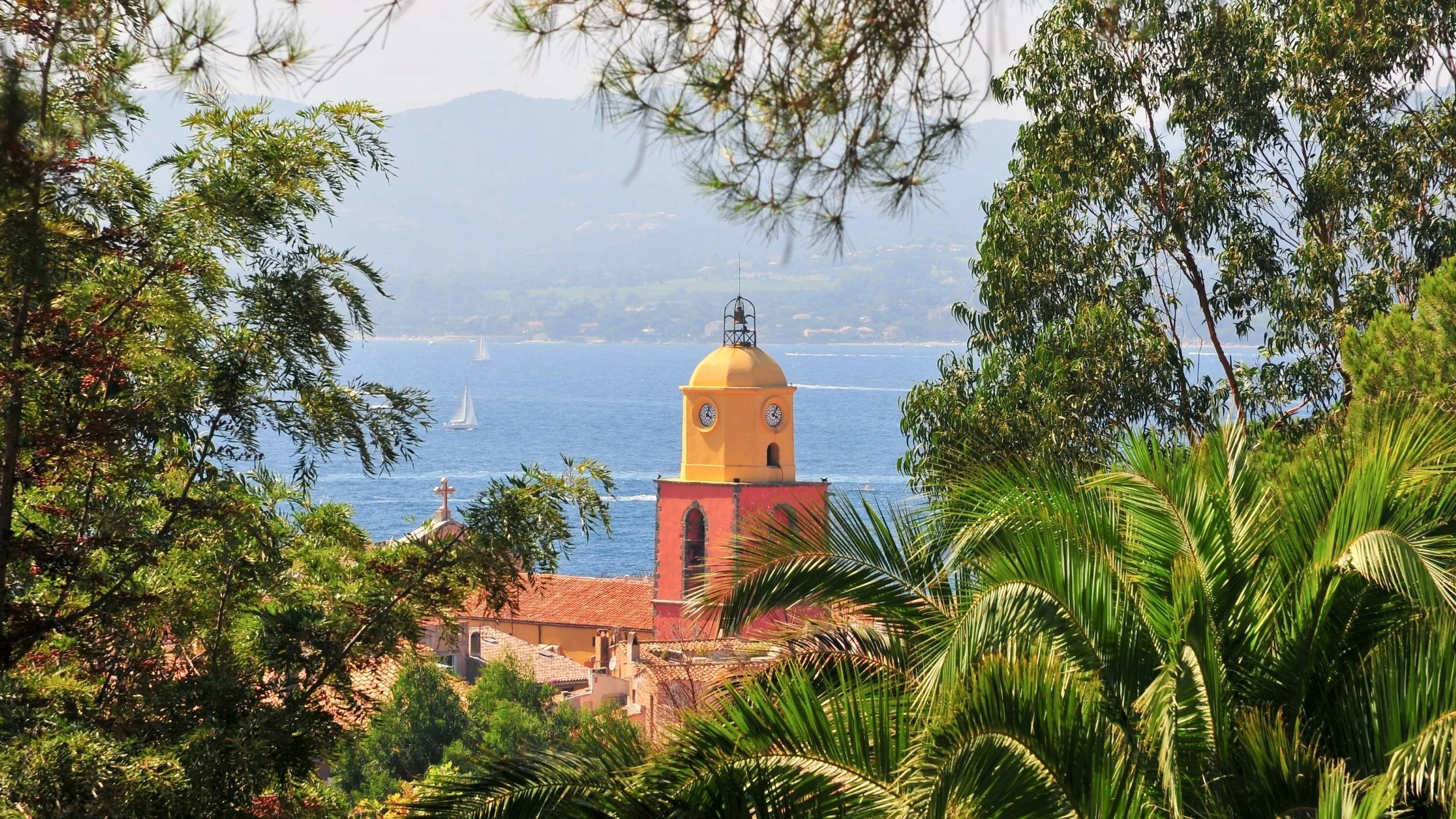 A view of a yellow clock tower with a red base, surrounded by lush green trees. The blue sea with sailboats and distant mountains are seen in the background.