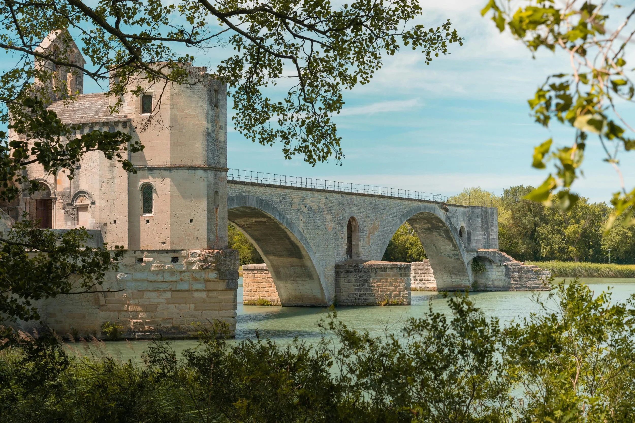 Historic stone bridge over a river, with a castle-like structure on one end, surrounded by trees and blue sky.