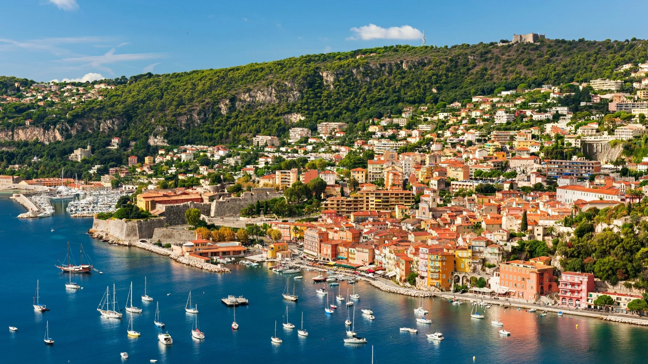 Scenic view of a coastal city with colorful buildings on a hillside overlooking a harbor filled with sailboats and yachts, with green hills and a clear blue sky in the background.