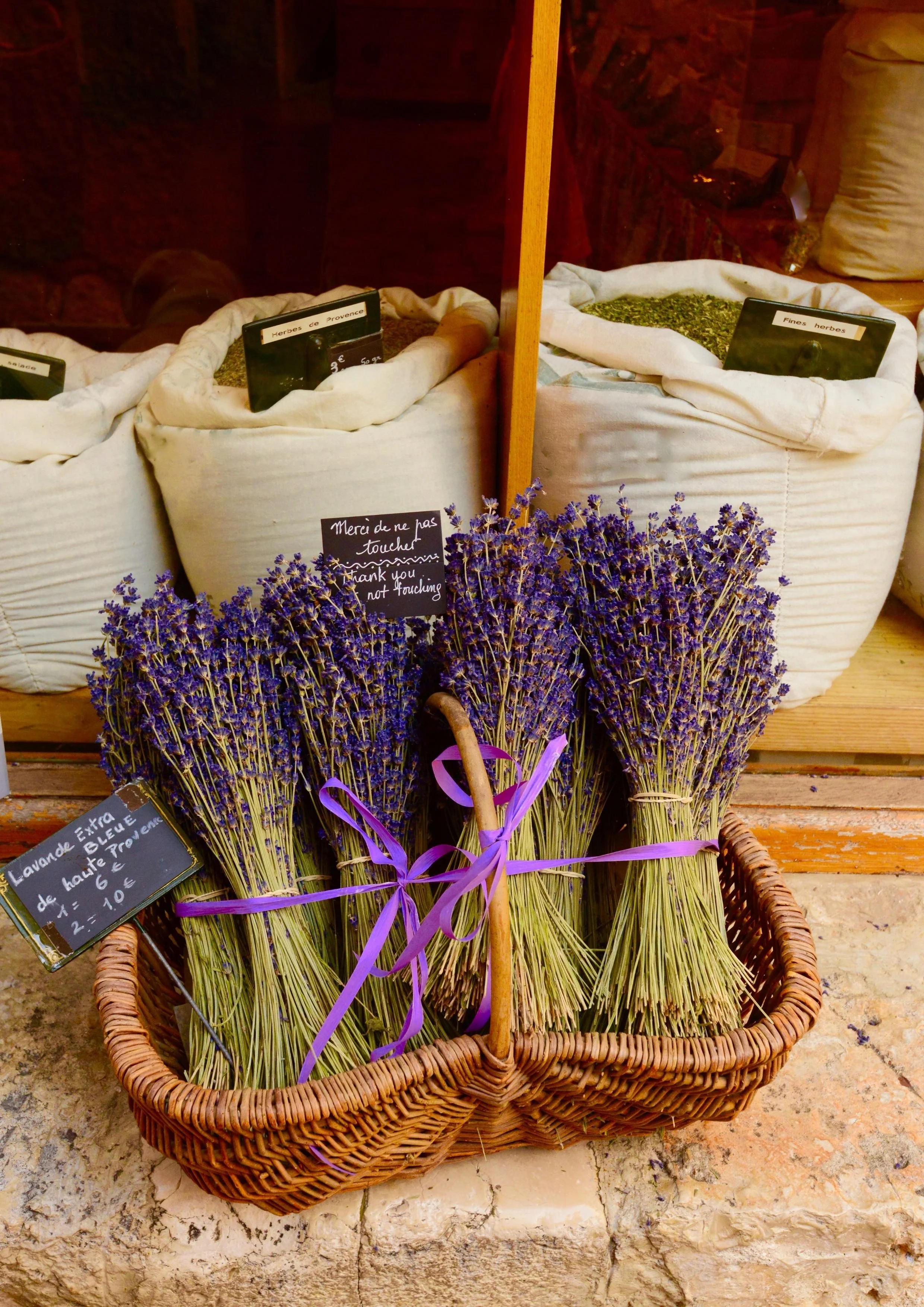 Bottes de lavande fraîche liées avec des rubans violets dans un panier en osier, avec un panneau indiquant 'Lavande d'exception de haute Provence', à côté de sacs d'herbes séchées et d'autres herbes dans une devanture de boutique.