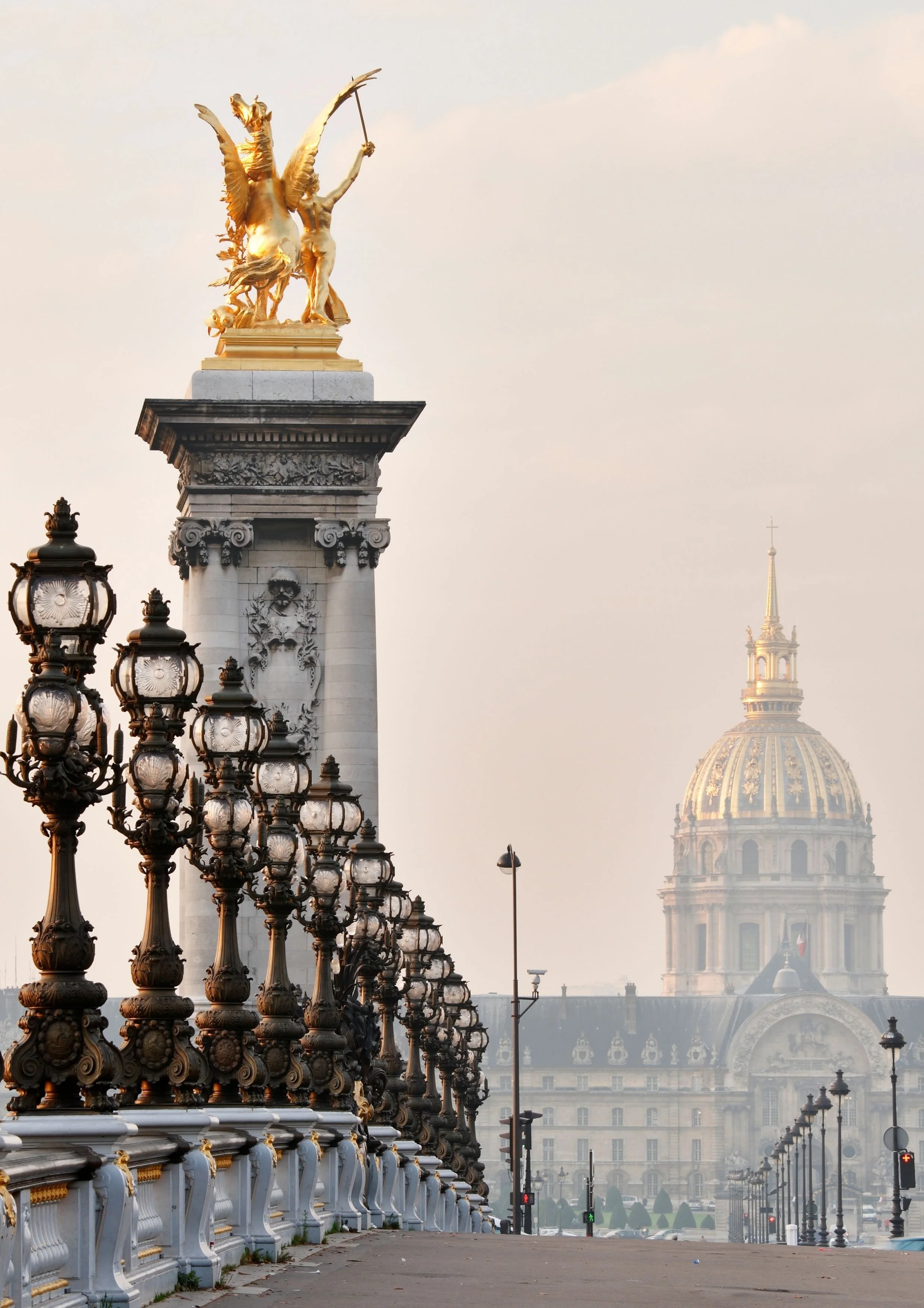 View of a bridge with ornate street lamps and statues at the top of a column, leading to a historic building with a large dome in the background.