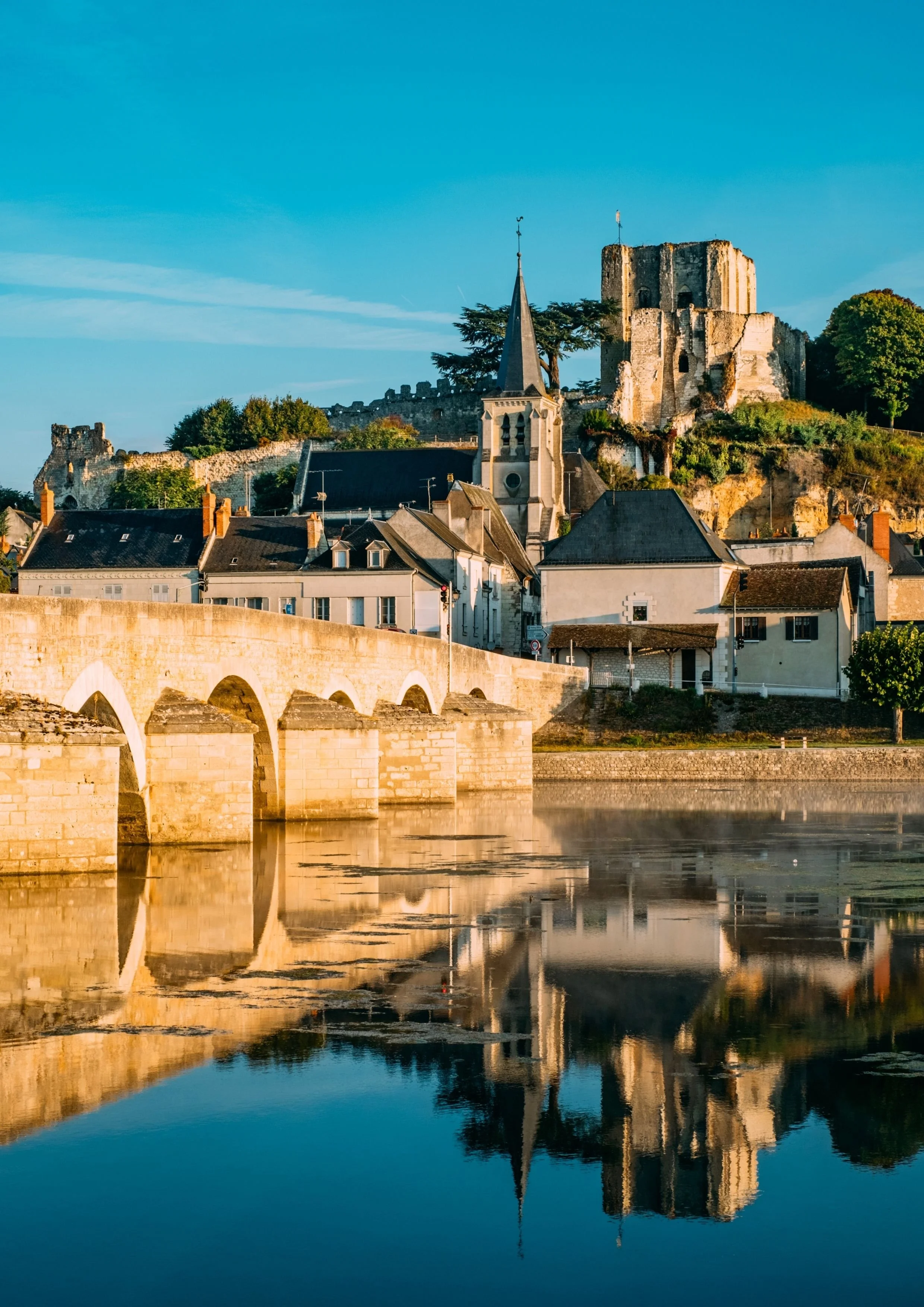 Une ville avec un château en ruines au sommet d'une colline, une église avec un clocher pointu et un pont ancien sur une rivière, le tout reflété dans l'eau, au coucher du soleil.