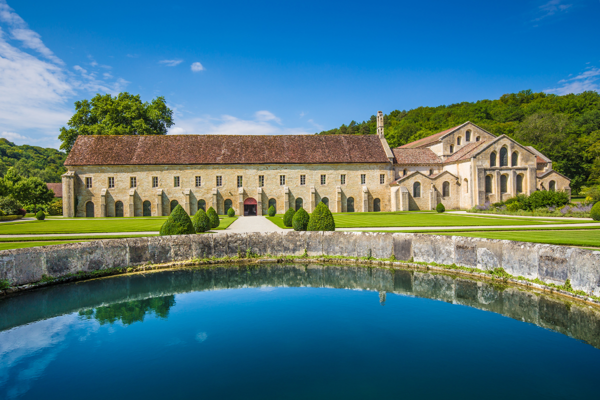 A historic stone church with a red-tiled roof, surrounded by green lawns, landscaped bushes, and a reflective pond in the foreground under a bright blue sky.