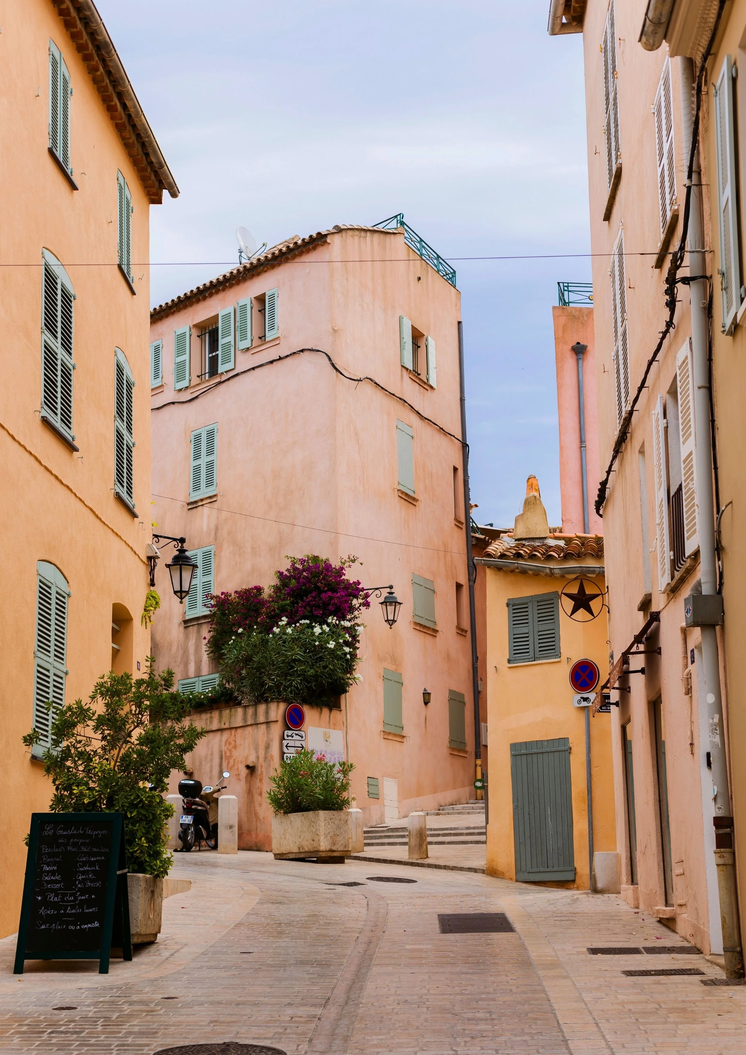 Narrow European street with pastel-colored buildings, closed shutters, potted plants, a scooter, and street signs.