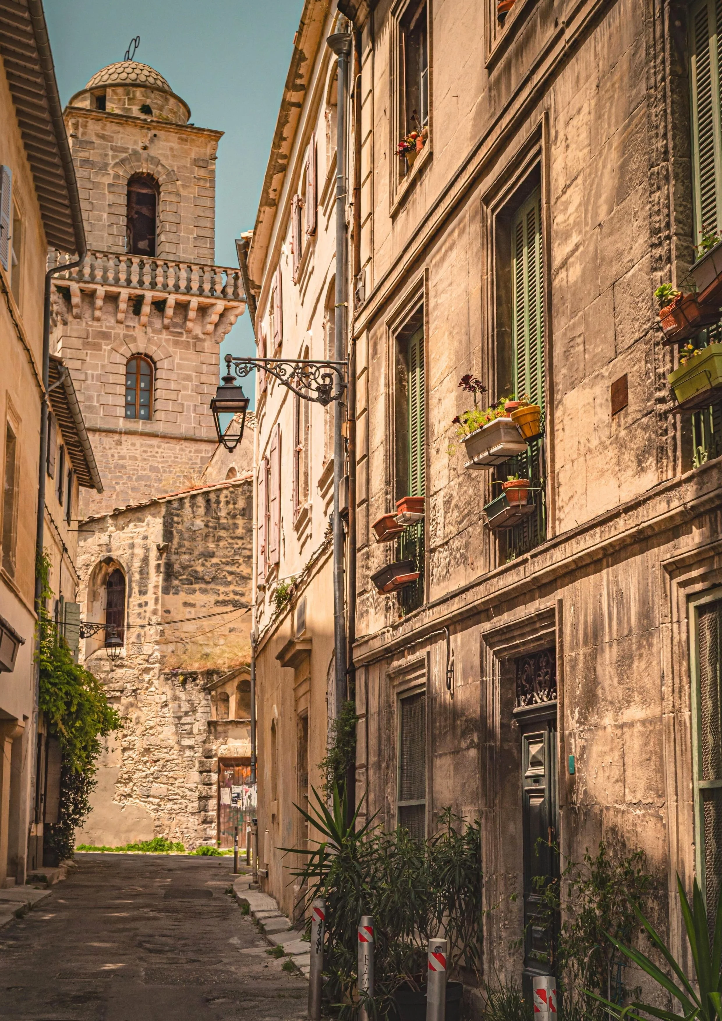 Une ruelle pavée bordée de vieux immeubles en pierre avec des fenêtres ornées de jardinières et une tour en arrière-plan. Le ciel est clair et ensoleillé.