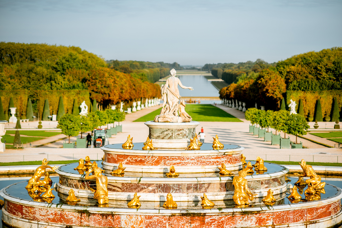 Une fontaine ornée de sculptures dorées représentant des personnages mythologiques dans un jardin bien entretenu avec des arbres aux feuilles colorées, un canal ou un plan d'eau en arrière-plan et un ciel partiellement nuageux.