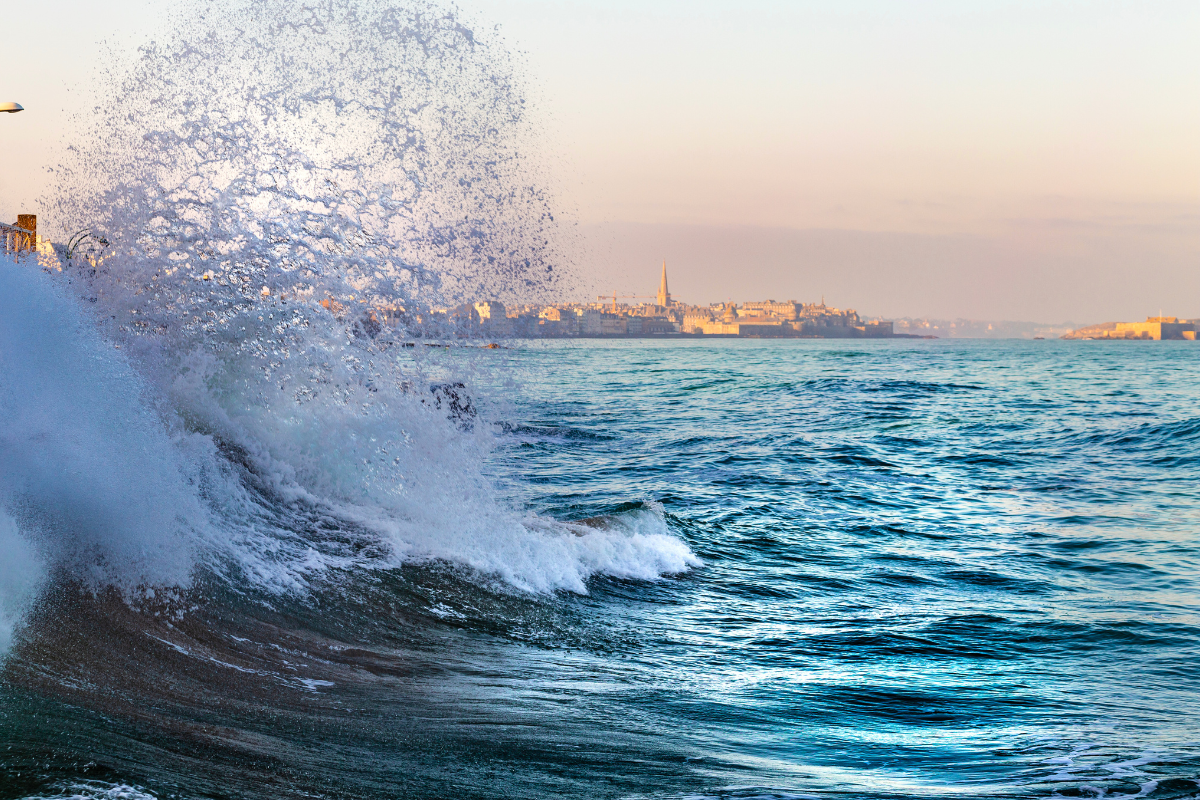 Vague de mer avec paysage urbain en arrière-plan, y compris une église ou un monument pointu, lors du coucher du soleil.
