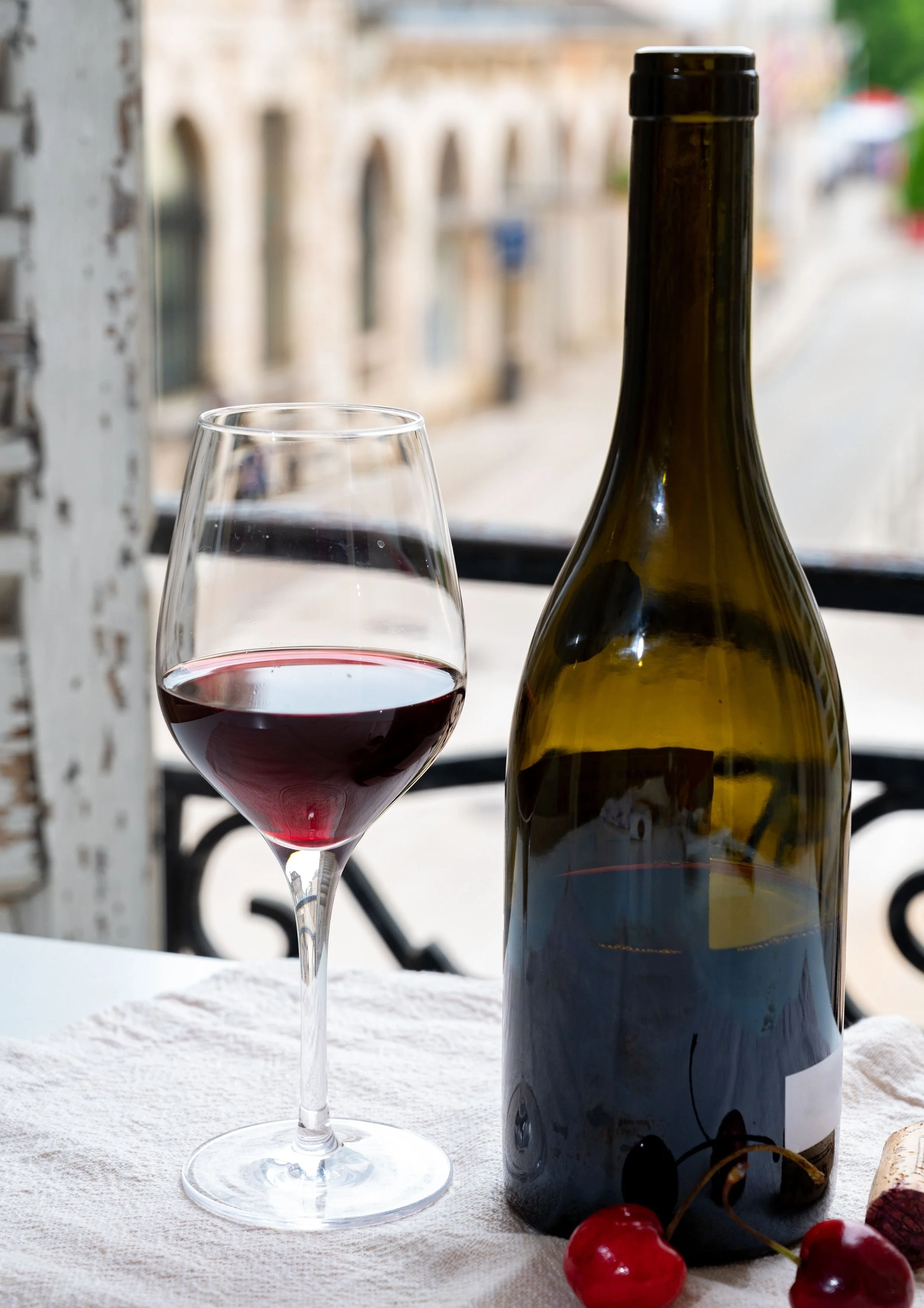 Une bouteille de vin rouge et un verre de vin rouge sur une nappe blanche, avec des cerises, à côté d'une fenêtre avec vue sur la ville