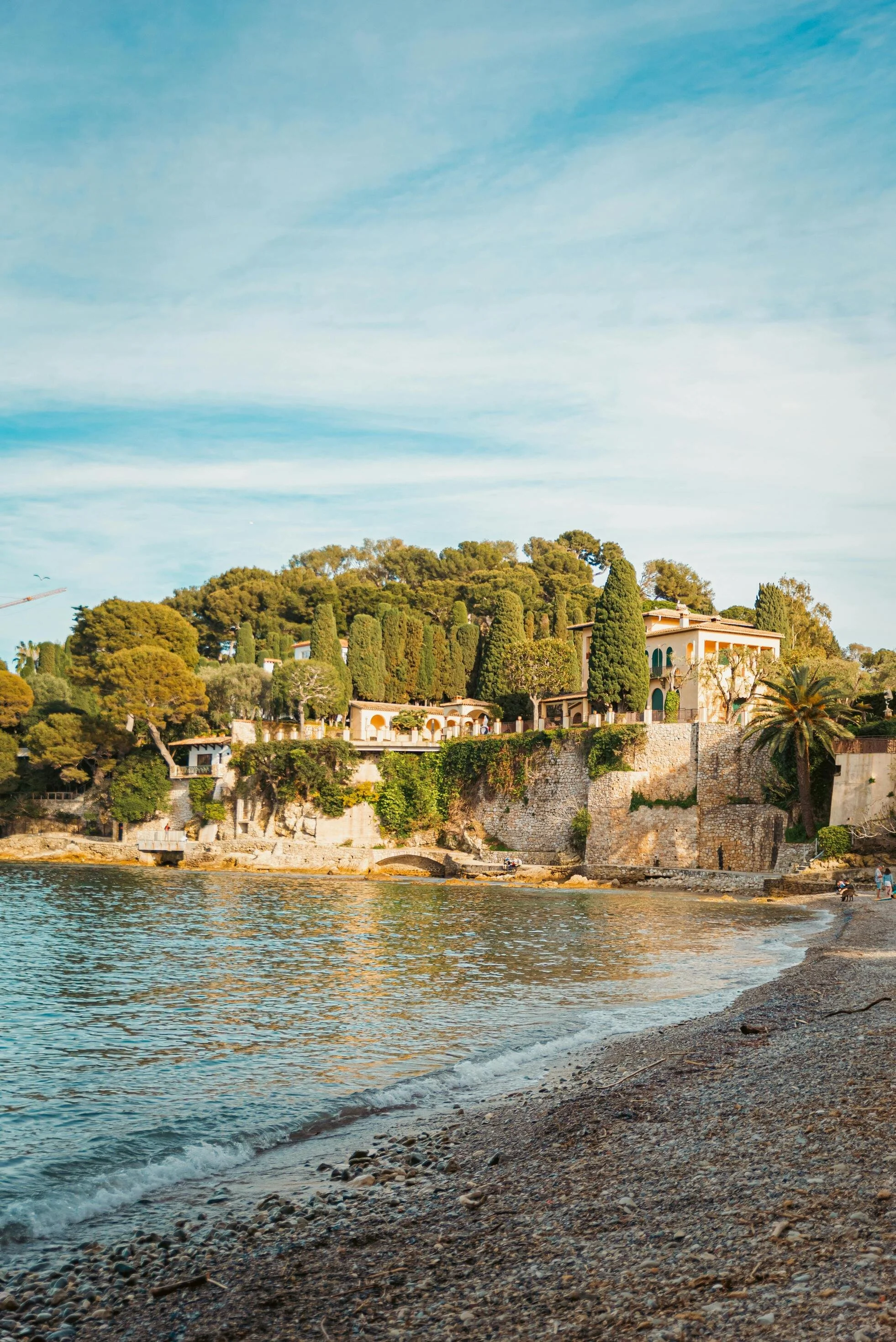 View of a scenic coastline with a pebble beach, calm water, and luxurious hillside homes surrounded by lush green trees.