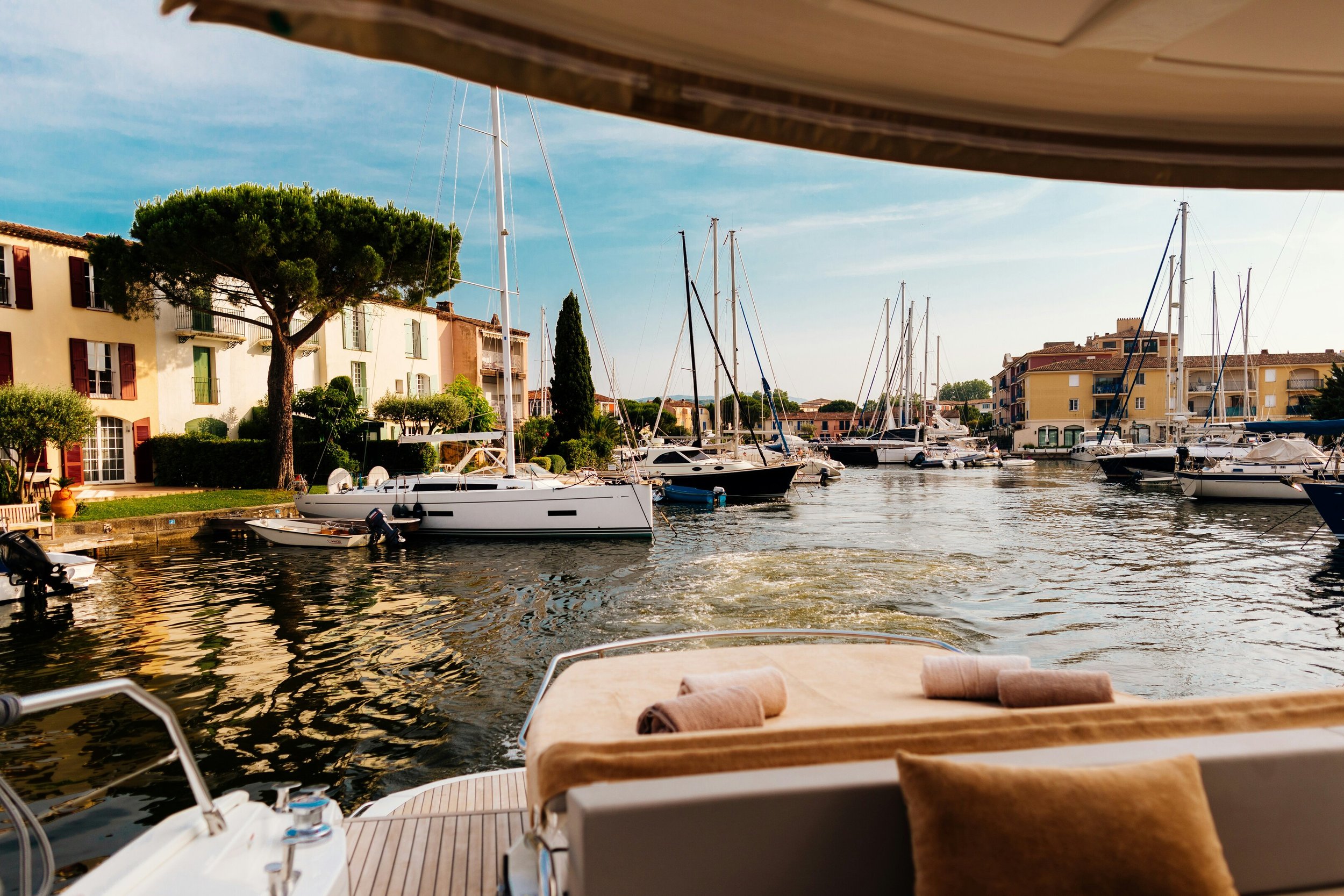 View from a boat looking out over a marina with sailboats docked, colorful buildings along the shore, trees, and a partly cloudy sky.