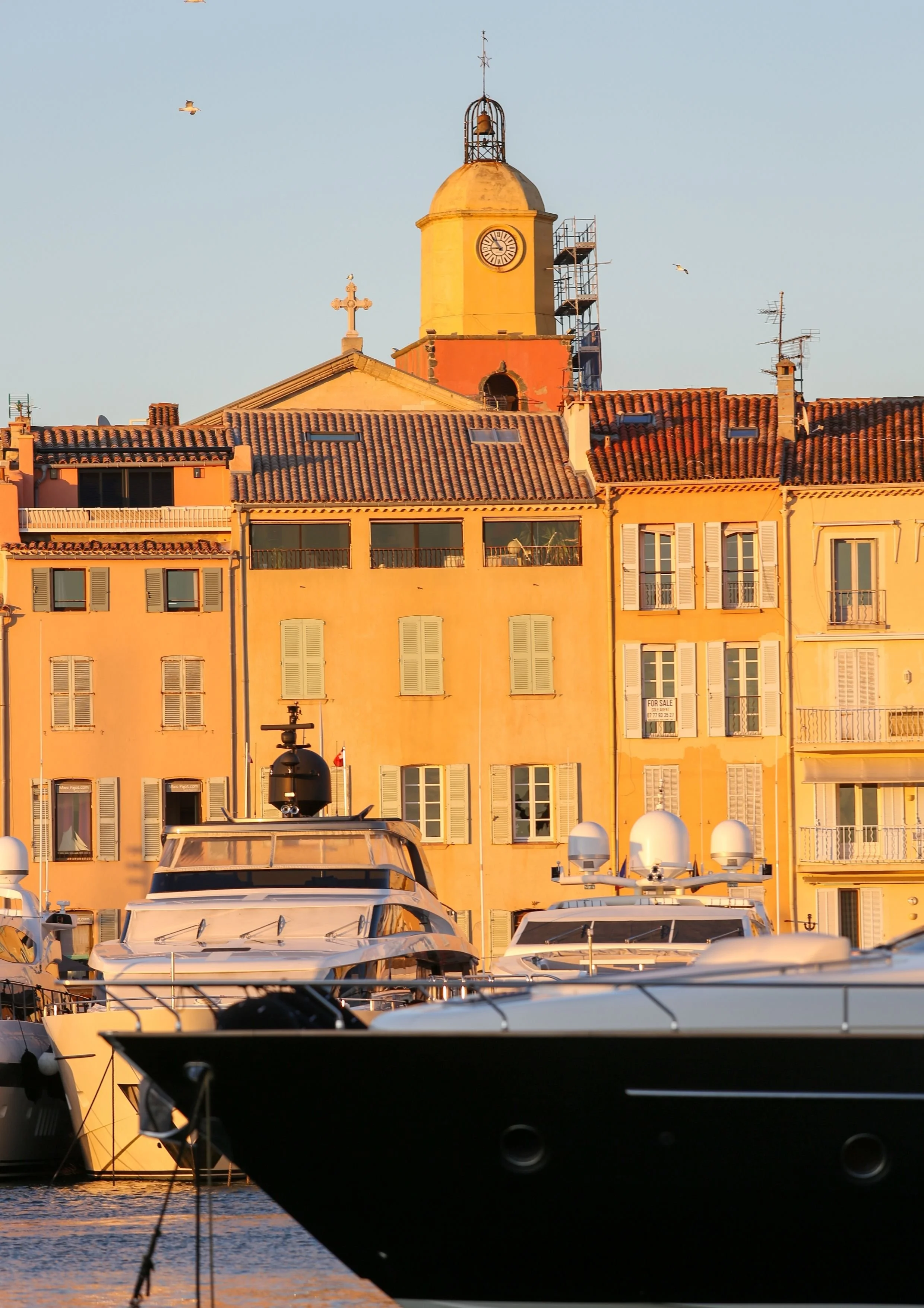 Port de plaisance avec des yachts au premier plan et des bâtiments colorés en arrière-plan, dont une église avec un clocher, lors du coucher du soleil.