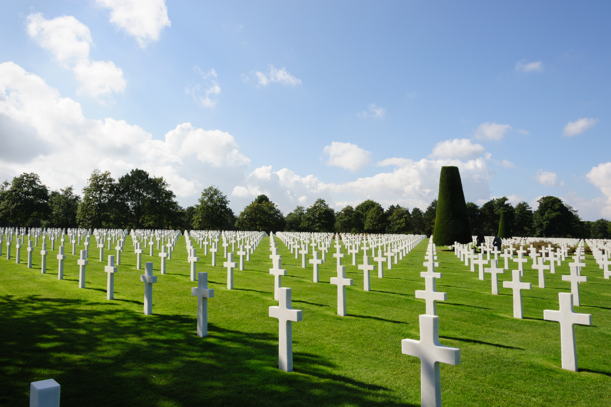 A military cemetery with rows of white crosses on lush green grass, tall trees in the background, and a partly cloudy sky.