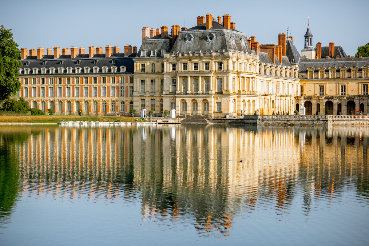 A grand historic castle with multiple chimneys and ornate architecture reflects in a large pond in front of it, with a peaceful landscape and clear sky.