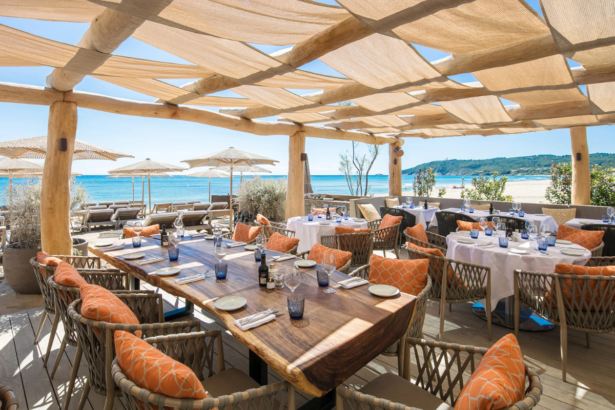 Restaurant en plein air avec vue sur la mer, tables élégantes avec napperons blancs, verres à vin bleus, assiettes blanches, et coussins orange sur des chaises en osier, sous un toit en tissus beige
