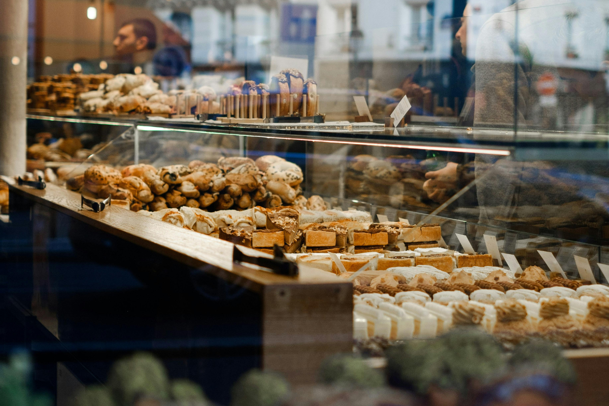 Vitrine d'une pâtisserie avec une variété de desserts, notamment des éclairs, des cookies, des petits fours et des tartes, en vitrine, avec reflection des passants et bâtiments extérieurs.
