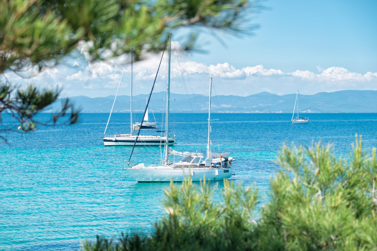 Sailboats anchored in turquoise water near a coastline with mountains in the background, framed by green foliage in the foreground.