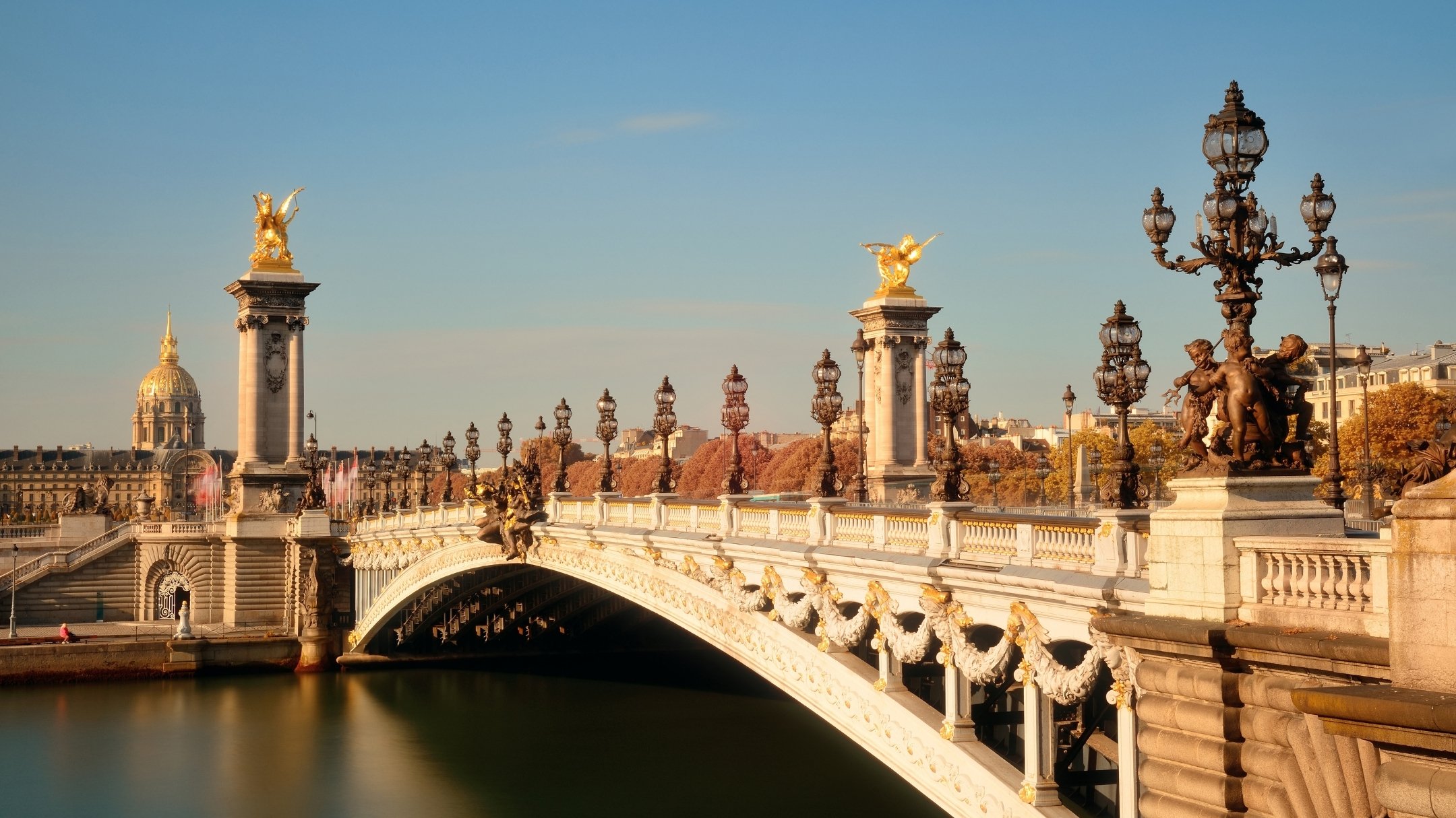 View of the Alexandre III Bridge in Paris, France, with ornate street lamps, gilded sculptures, and historic buildings in the background.