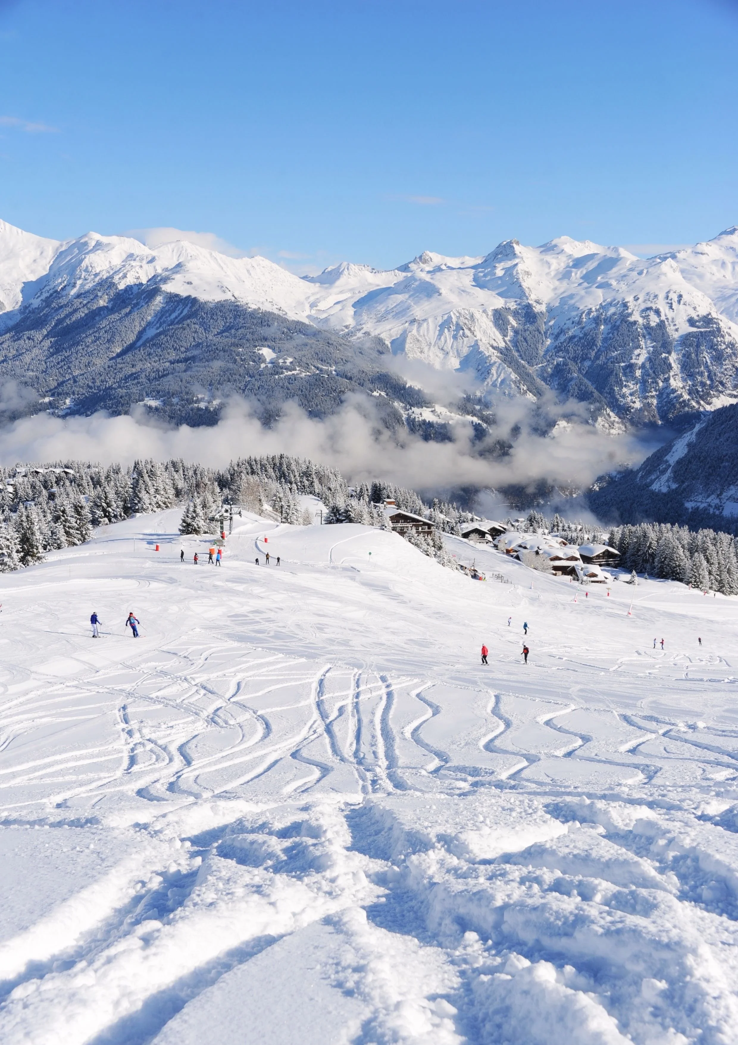 Piste de ski enneigée avec des skieurs, fond de montagnes enneigées sous un ciel bleu.