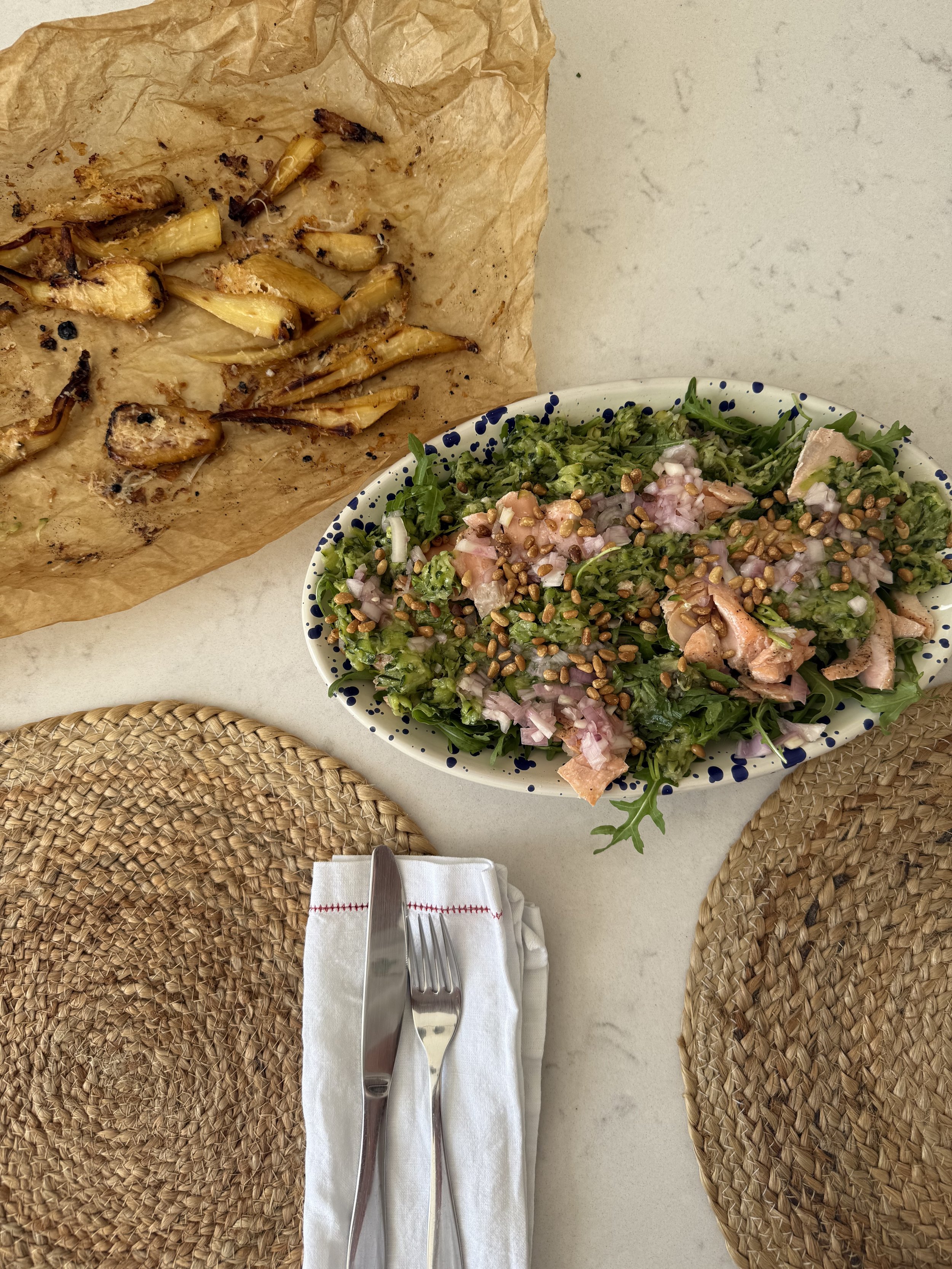 A plate of fresh green salad with chopped vegetables and seeds, a paper-lined tray of roasted small vegetables or early harvested root vegetables, and two woven placemats with a fork and knife on a napkin, on a white table.