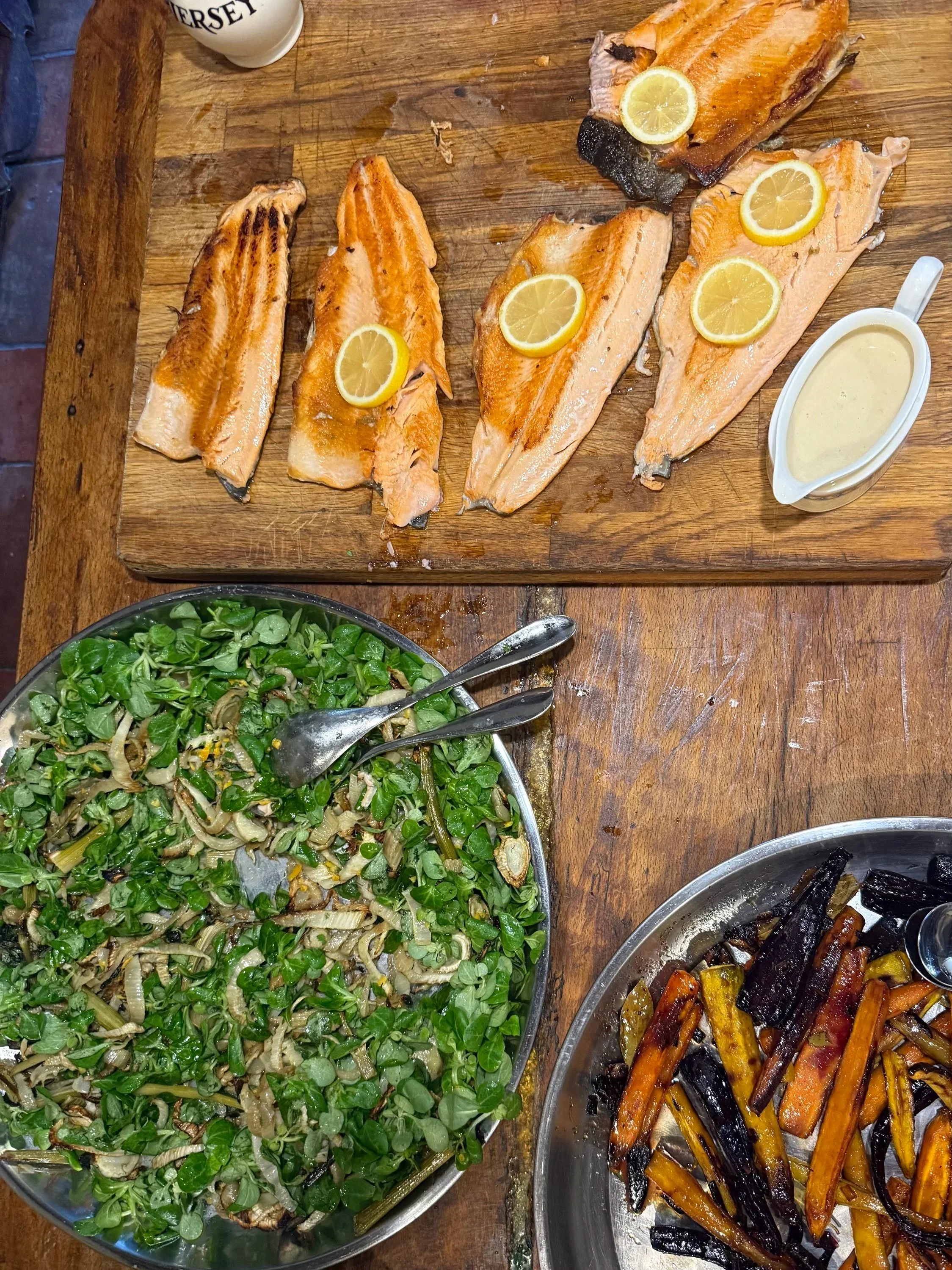 Grilled salmon fillets with lemon slices and tartar sauce, a bowl of sautéed greens with onions, and roasted carrots with eggplant on a wooden table.