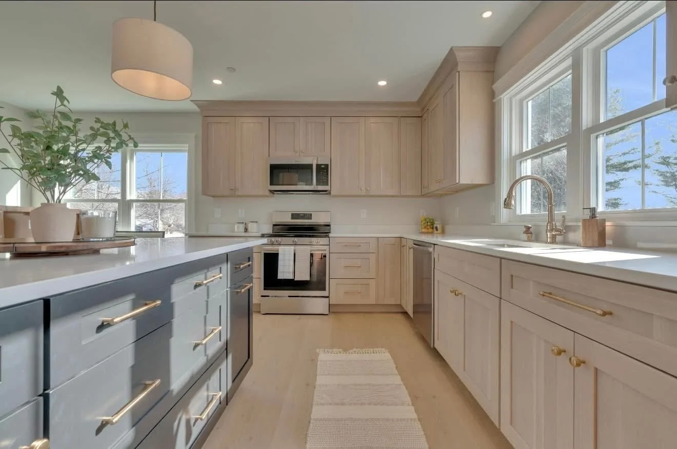 Bright kitchen with white and light wood cabinets, stainless steel appliances, a large window over the sink, and a light-colored kitchen island with gold handles.