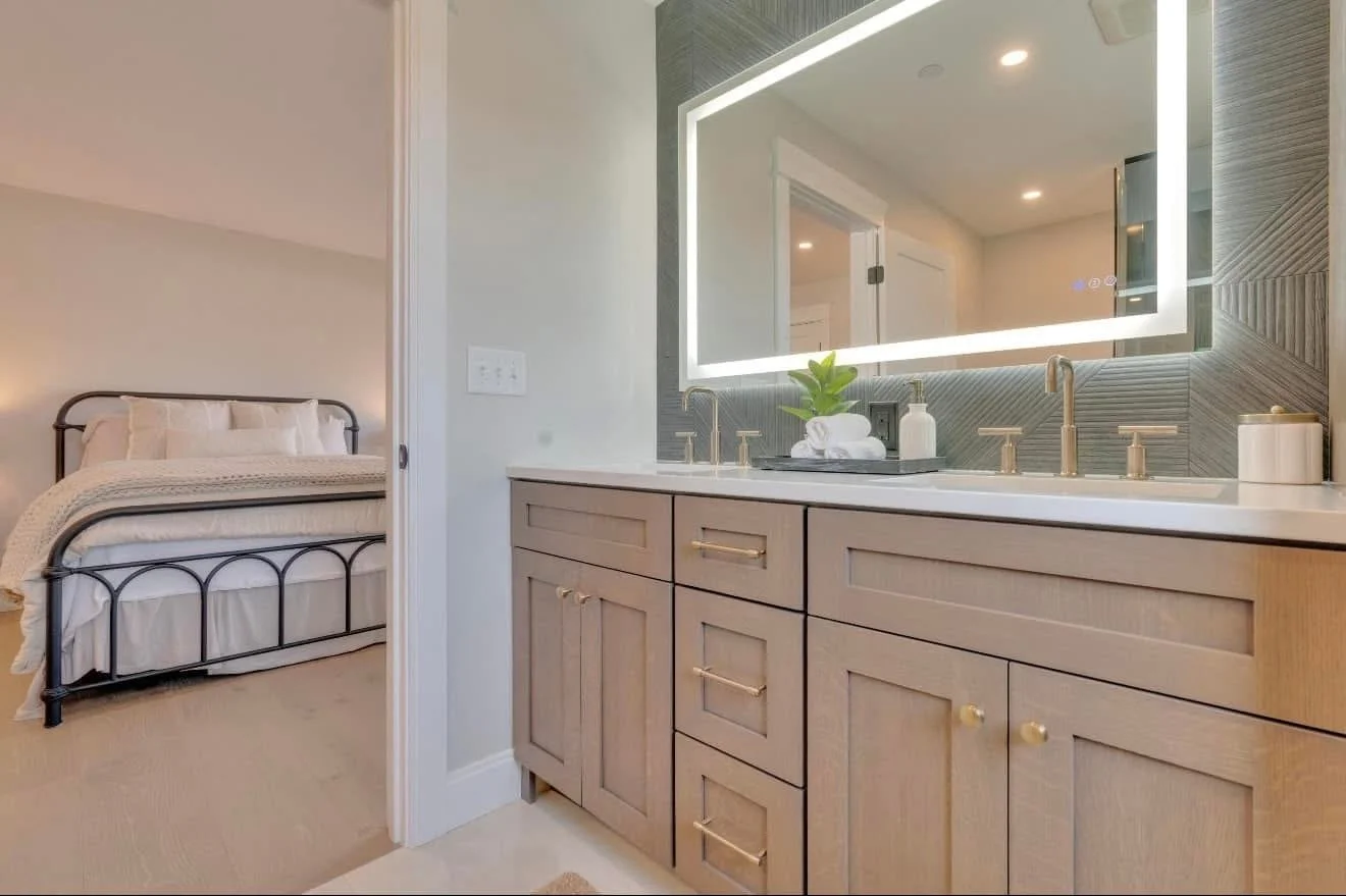 A bathroom vanity area with a large backlit mirror, a countertop with a small plant and toiletries, and a bedroom with a metal bed frame and bedding visible through an open door.