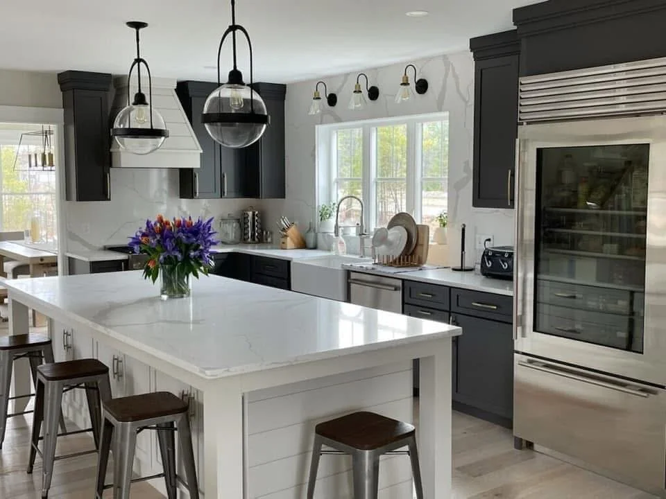 Modern kitchen with dark cabinets, white countertops, a kitchen island with a bouquet of purple flowers, pendant lights, and a large stainless steel refrigerator.