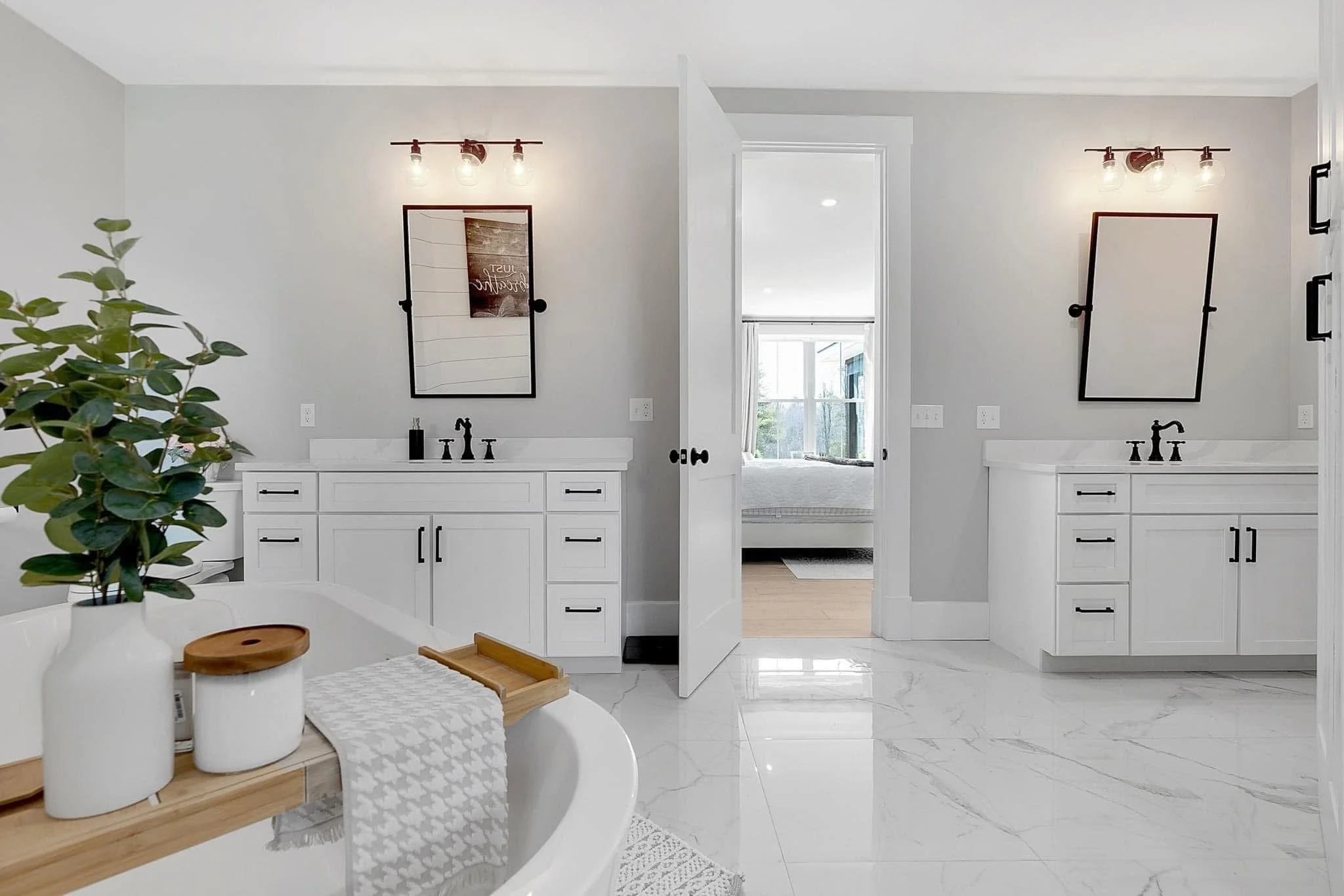 Bathroom with two white vanities, black fixtures, framed mirrors, and wall-mounted light fixtures, with a view into a bedroom through an open door, decorated in neutral tones and modern style.