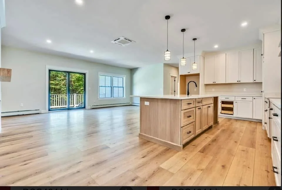Open-concept kitchen and living area with wood flooring, white cabinets, island with wooden cabinets, pendant lights, and large windows with a sliding door leading to a balcony.