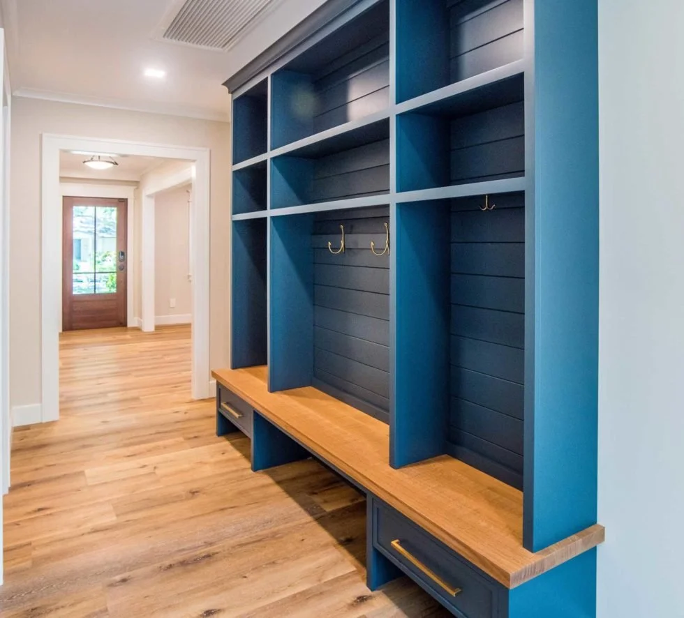 Blue built-in entryway storage with hooks for coats and shoes, located in a hallway with wood flooring and a front door in the background.