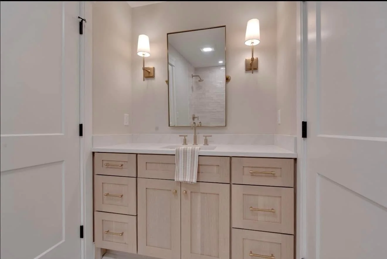 Bathroom vanity with a mirror, two wall-mounted sconces, a towel hanging from the faucet, wooden cabinets, and a white countertop.