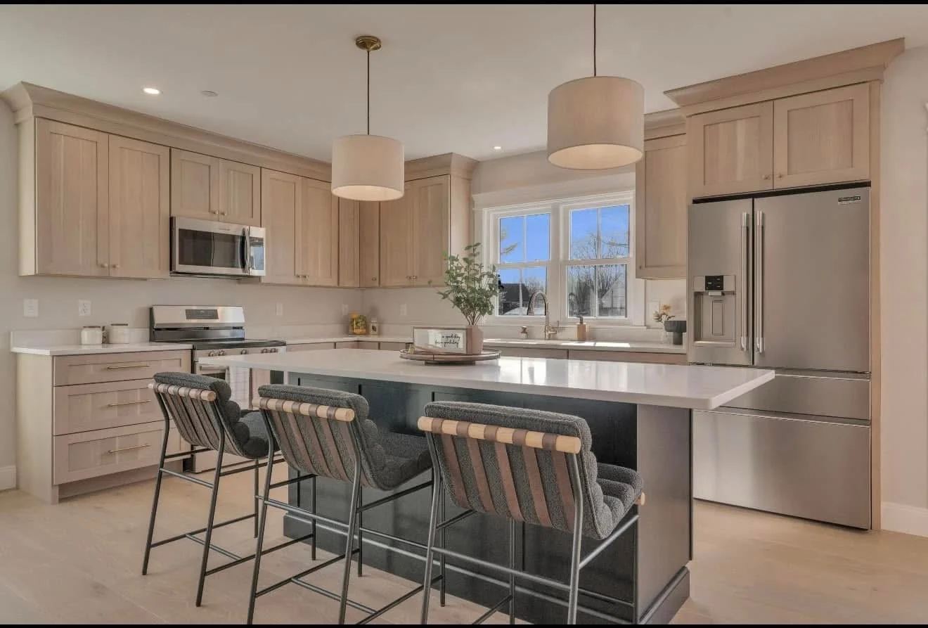 Modern kitchen with light wood cabinets, stainless steel appliances, and a central island with four upholstered bar stools. Large window behind the sink area letting in natural light.