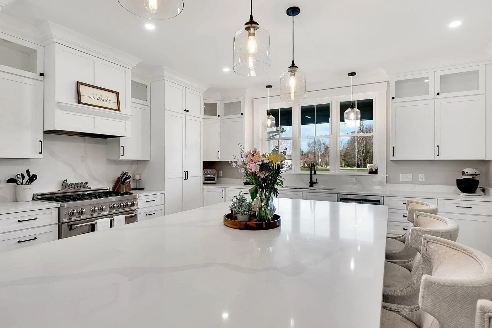 Modern white kitchen with large island, pendant lights, and window overlooking outdoors.