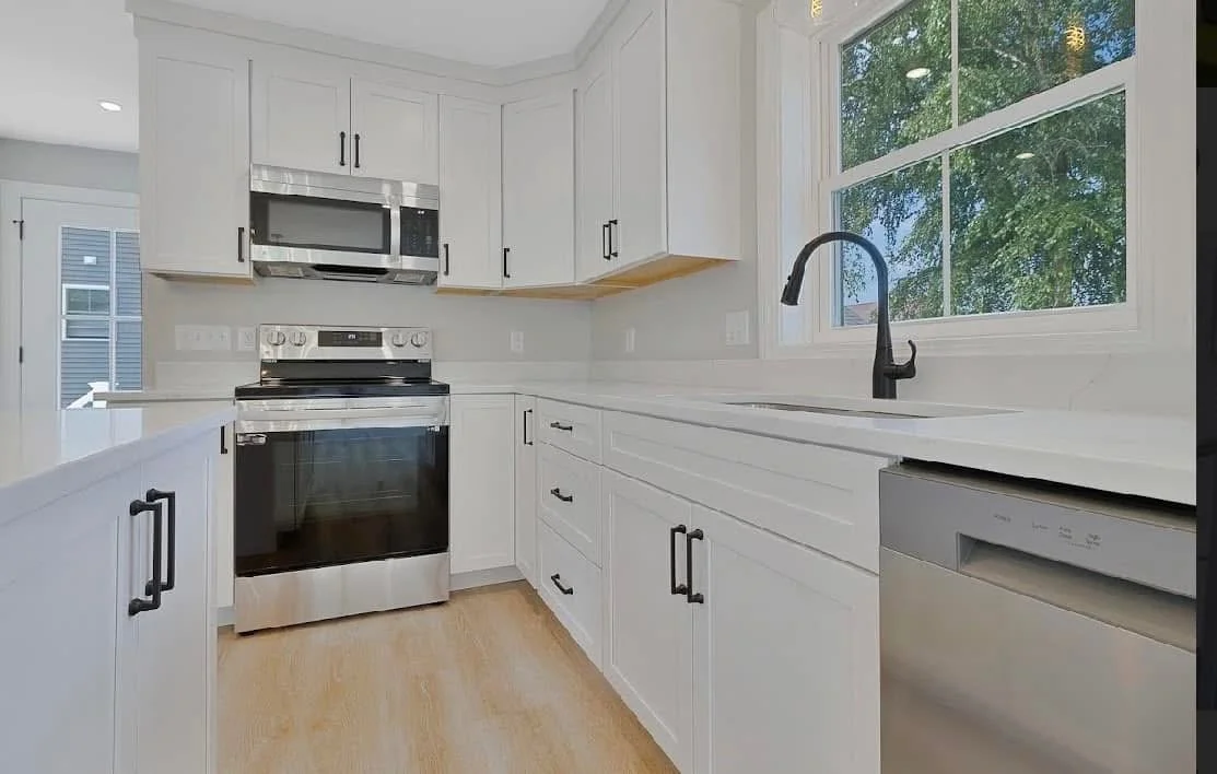 White kitchen with black hardware, stainless steel oven and microwave, black faucet, and light wood flooring, near a window overlooking trees.